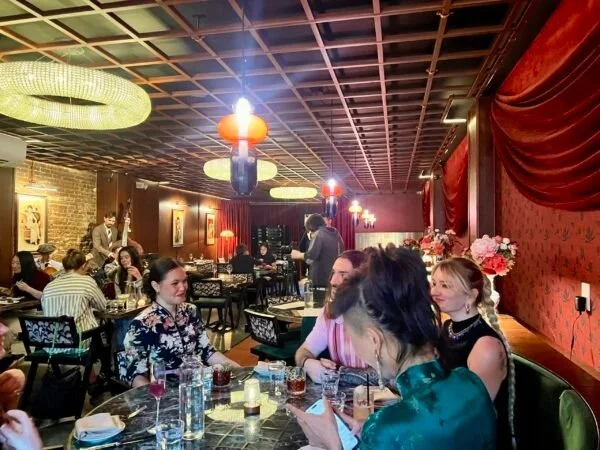 People dining in a dimly lit restaurant with red walls, decorated with flowers and artwork, featuring decorative hanging lights and a cozy atmosphere.