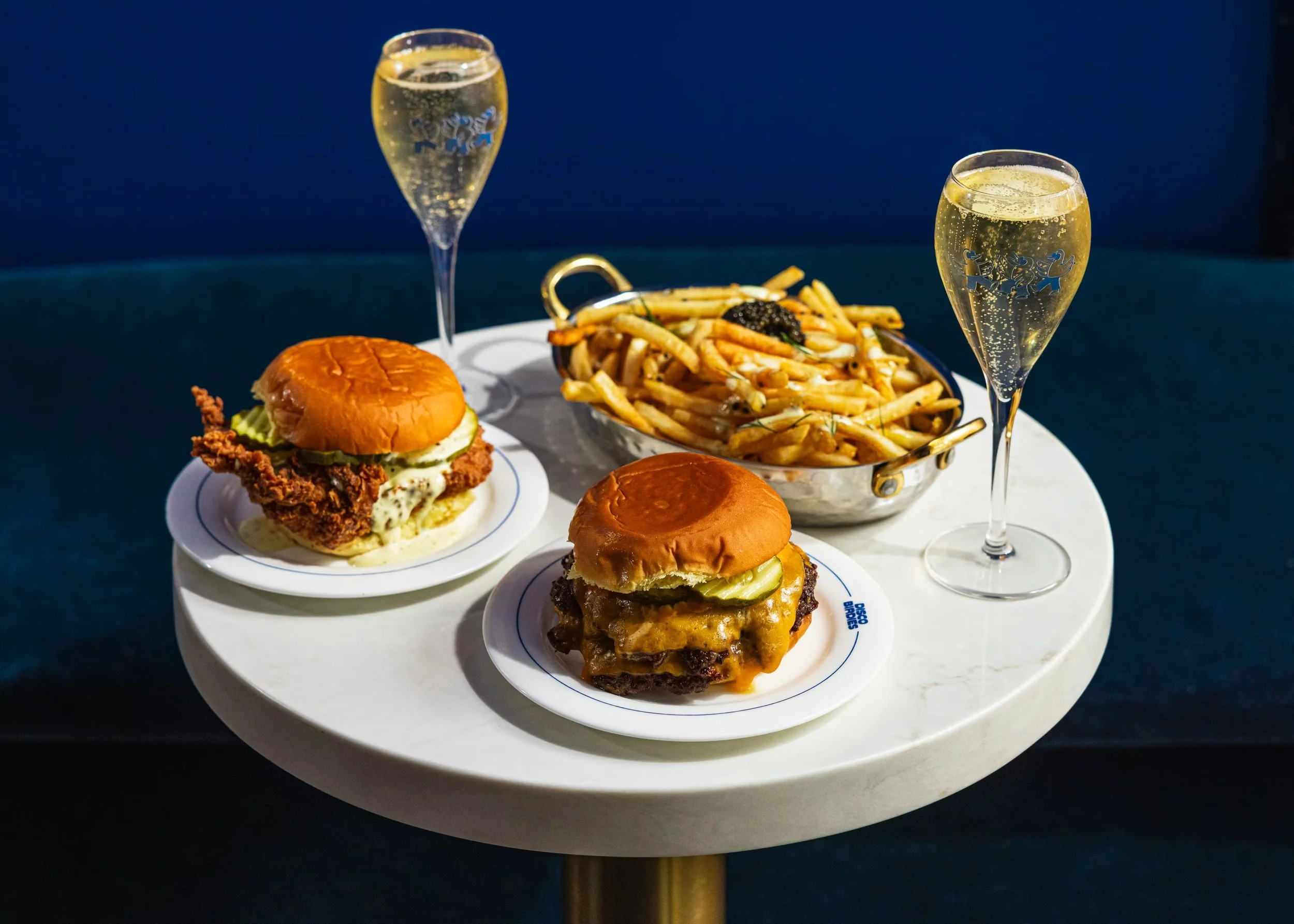Two cheeseburgers with pickles and cheese on small plates, a basket of French fries with black truffle garnishes, and two glasses of sparkling wine on a white round table with a dark background.