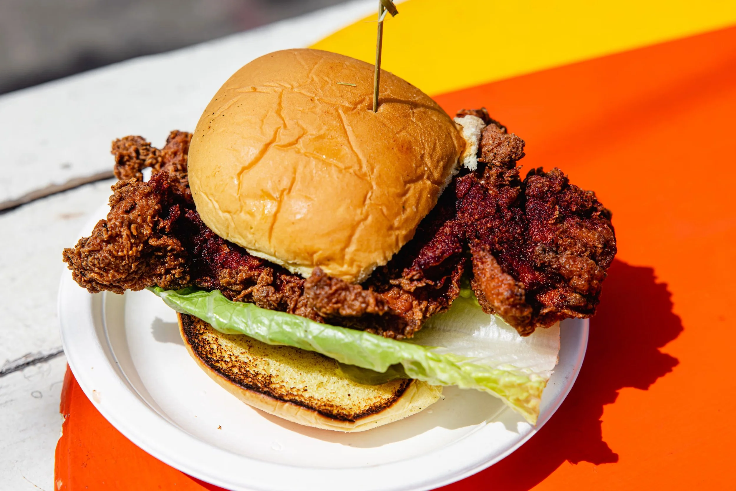 Fried chicken sandwich with lettuce, a bun, and a toothpick in a disposable white plate on an orange surface.