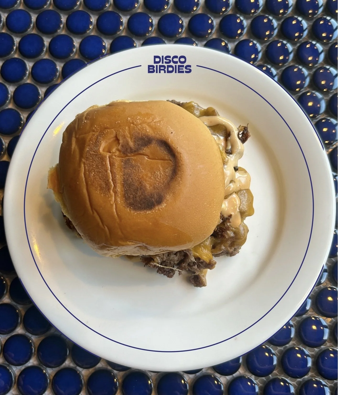 A cheeseburger with a toasted bun, melted cheese, cooked ground beef patty, and sauce, served on a "Disco Birdies" branded plate.