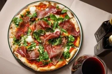 Pizza topped with prosciutto, arugula, and mozzarella, alongside a glass of red wine.