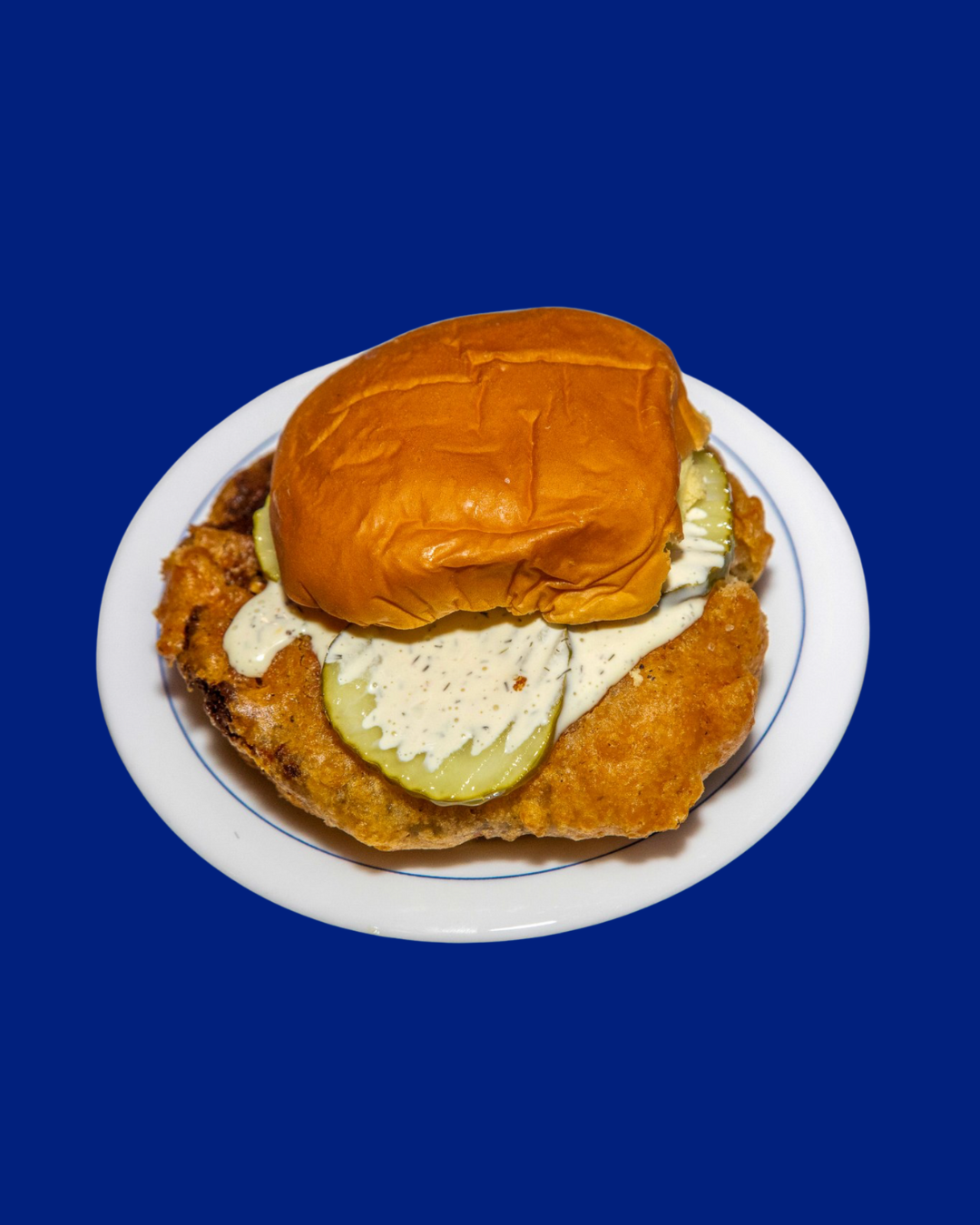 Fried chicken sandwich on a white plate with pickles, lettuce, mayonnaise, and a toasted bun against a blue background.