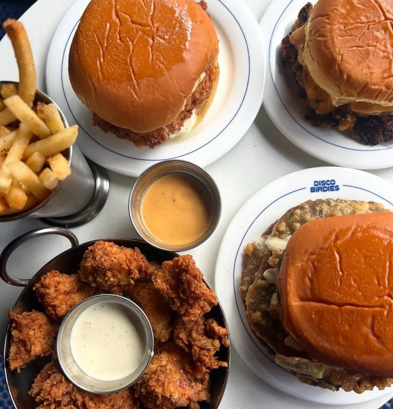 Three cheeseburgers with buns, two plates of fries, a bowl of fried chicken with gravy, and a cup of dipping sauce on a table.