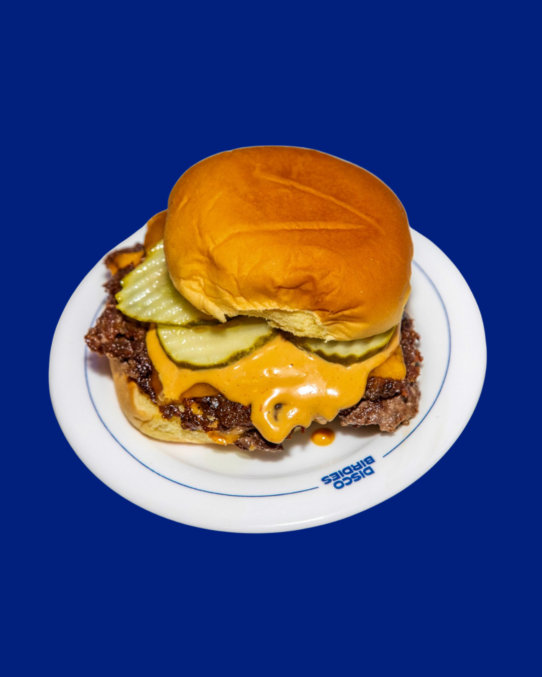 A cheeseburger with pickles on a white plate against a blue background.