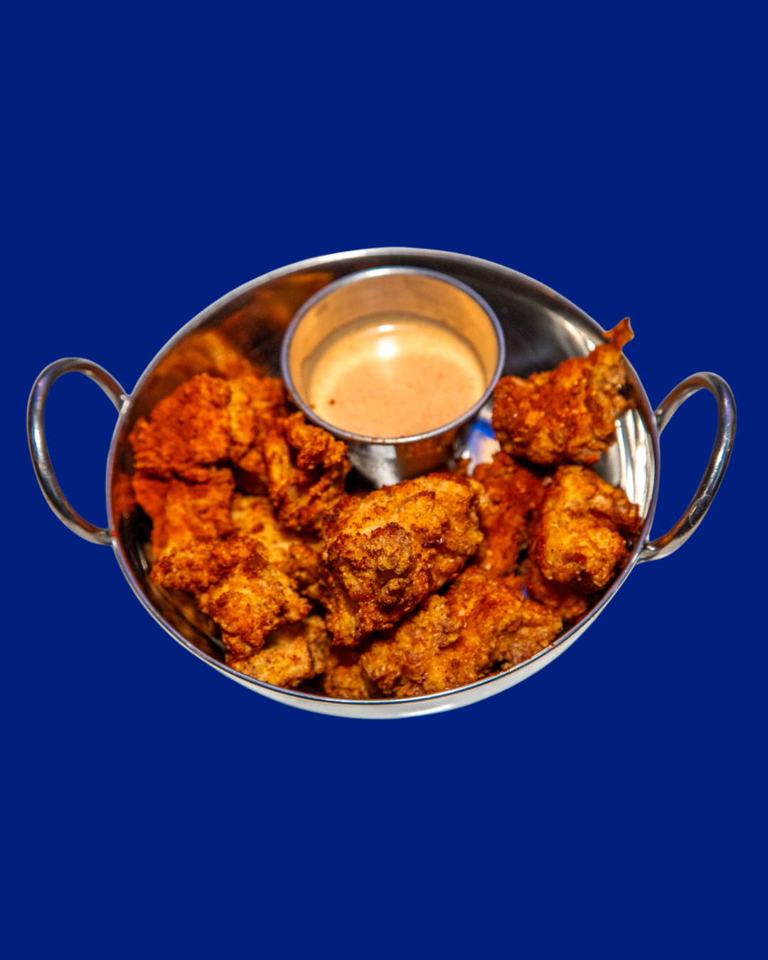 Fried chicken pieces with dipping sauce in a round metal serving tray with two handles against a dark blue background.