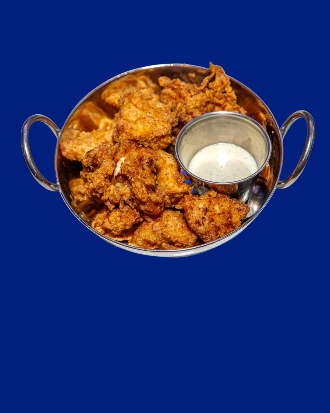 Basket of fried chicken pieces with a small cup of white dipping sauce against a solid blue background.