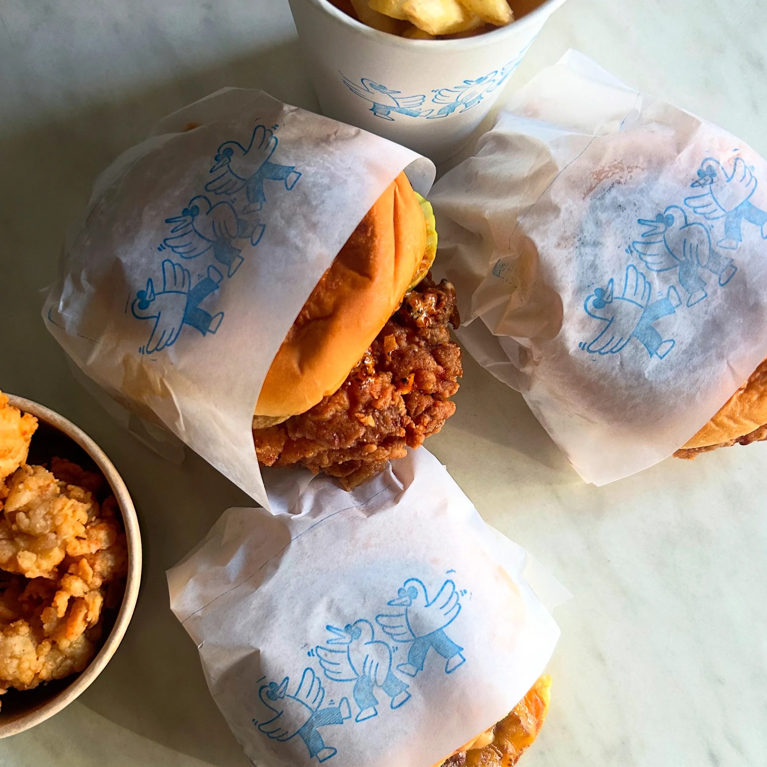 Two fried chicken sandwiches wrapped in white paper with a blue duck logo, alongside a bowl of fried chicken and a cup of fries.