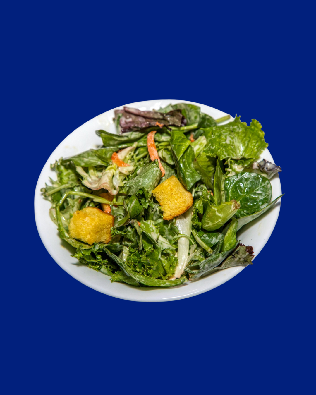 A white bowl filled with mixed green salad, including lettuce, spinach, and shredded carrots, with a couple of croutons on top, placed against a plain blue background.