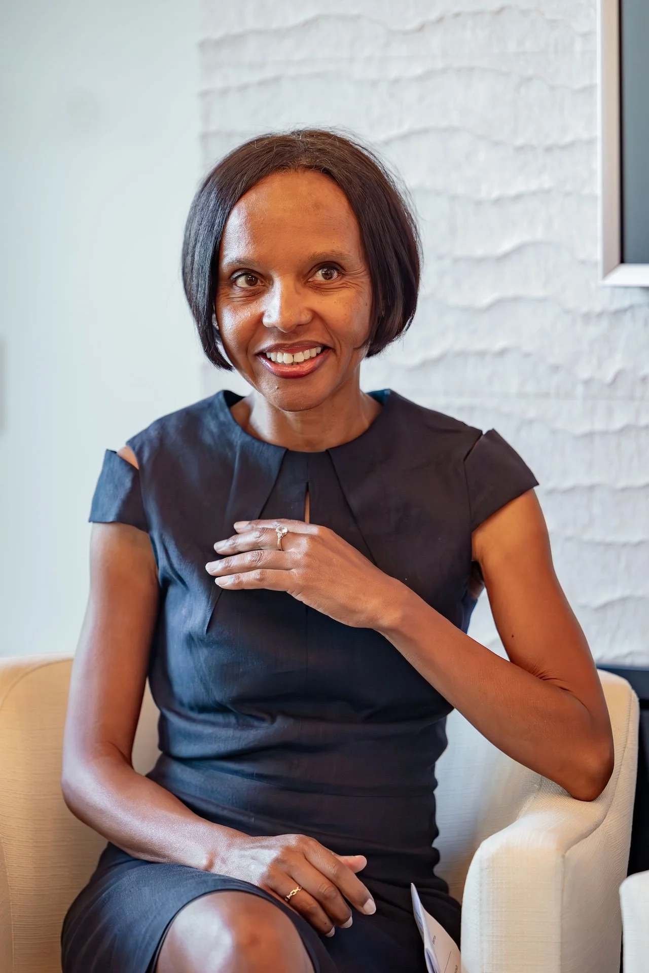 A woman with short dark hair, wearing a black dress, sitting on a beige chair, smiling and touching her chest with her right hand, with a white brick wall in the background.