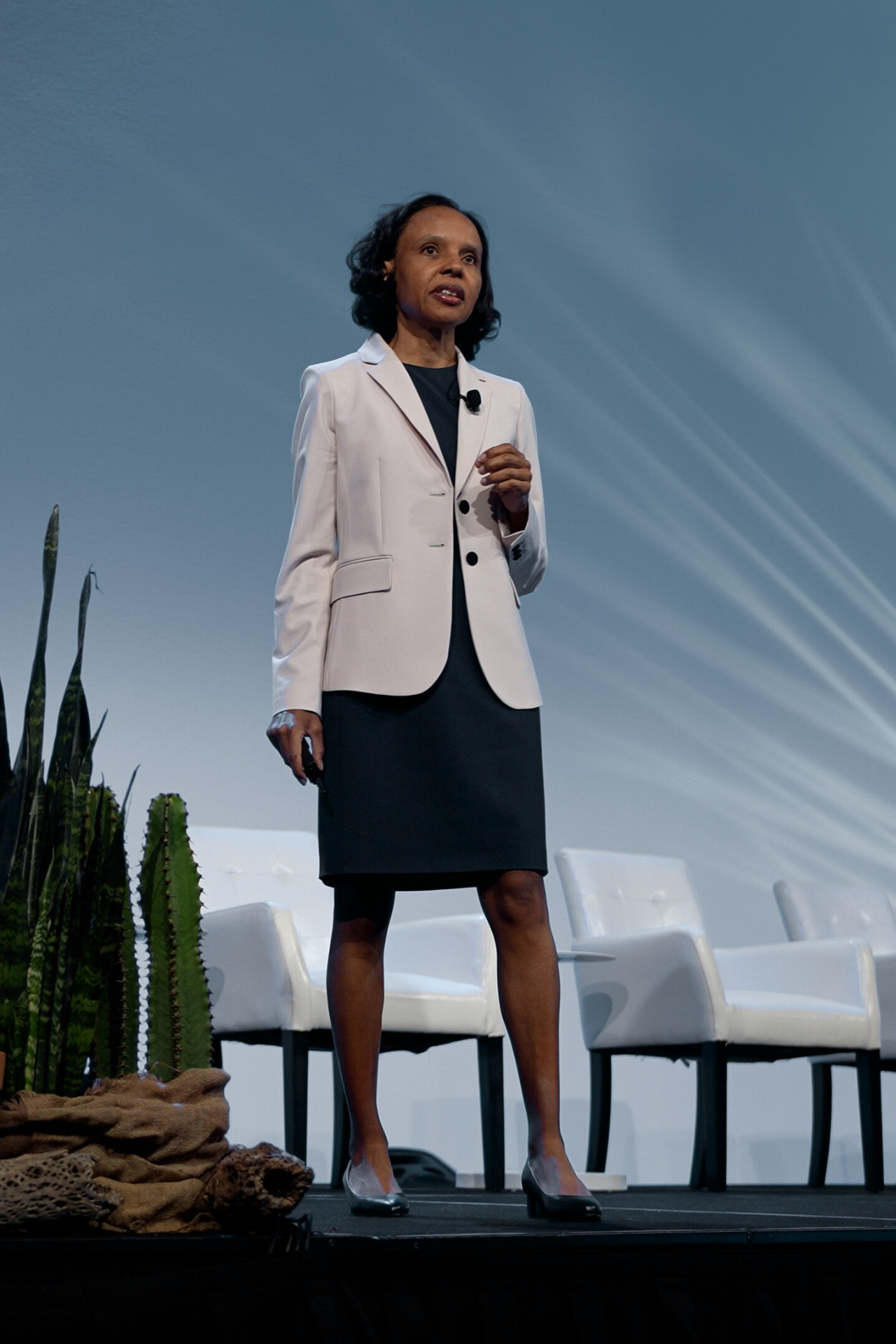 A woman standing on a stage with a microphone, wearing a white blazer and black dress, with potted cacti and white chairs in the background.