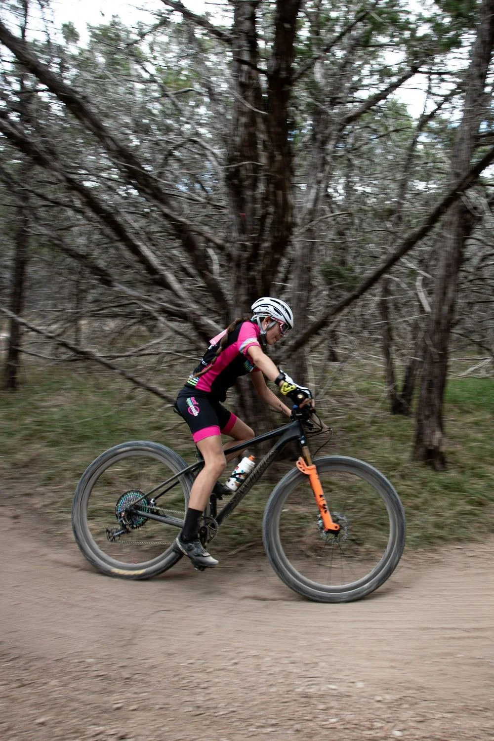 An Alamo City Femmes rider in a pink and black team kit riding a wooded dirt trail