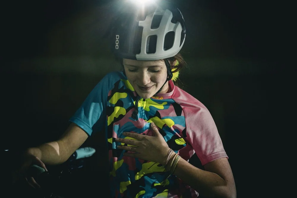 A rider in a bright team jersey and helmet light, smiling at dusk