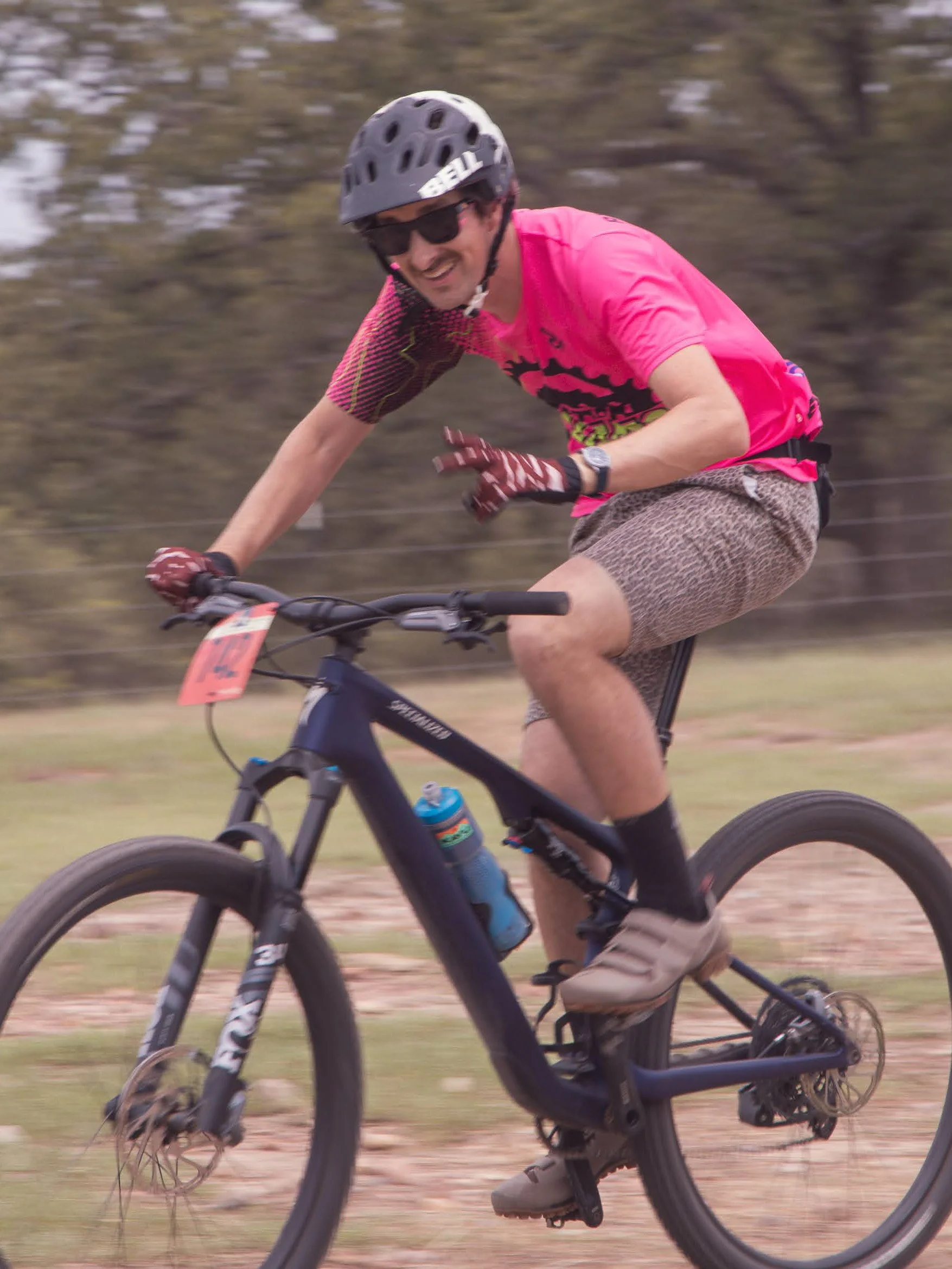 A man riding a mountain bike outdoors wearing a black helmet, sunglasses, pink shirt, patterned shorts, and gloves, smiling and enjoying the ride.