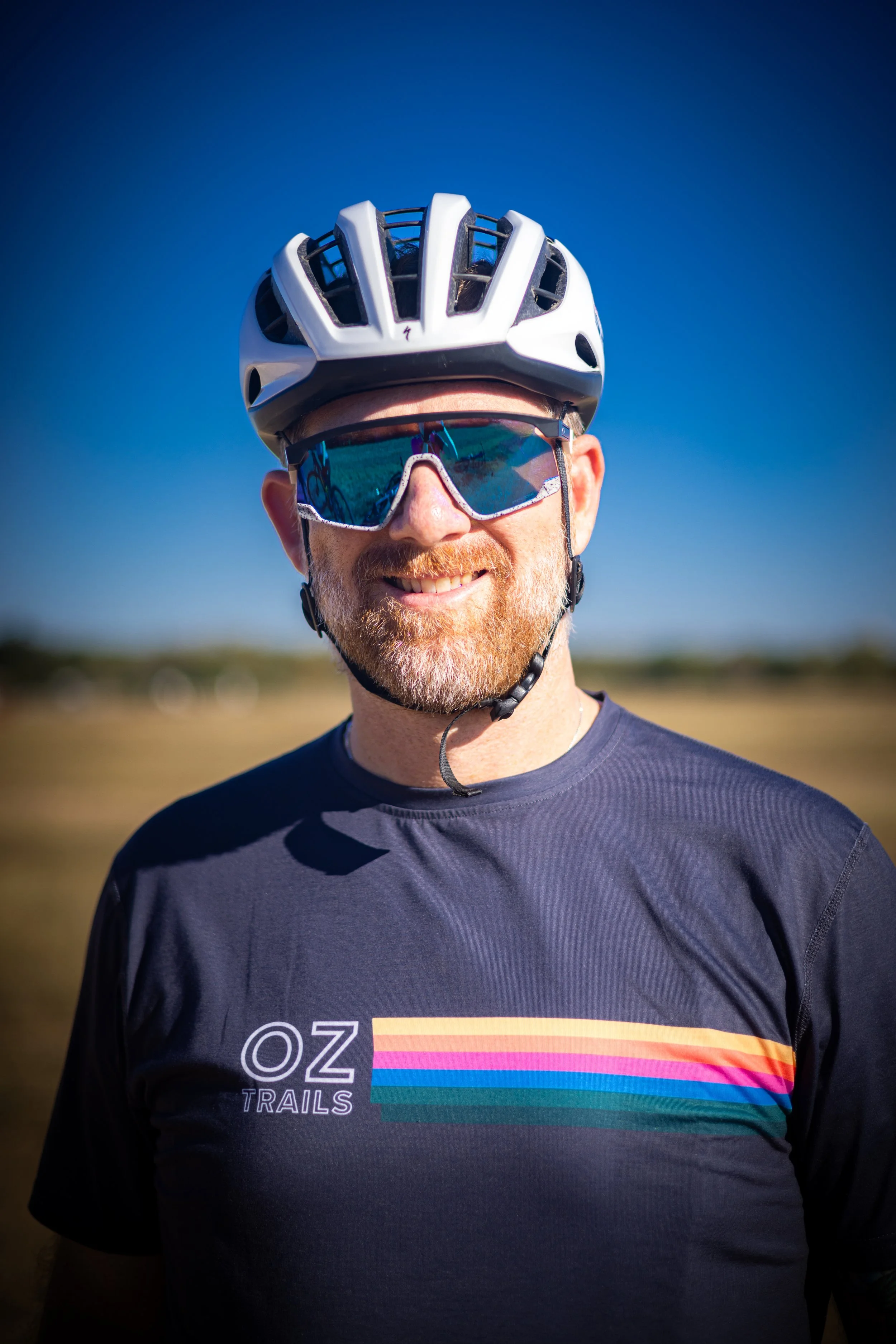 Mountain biker wearing a helmet, sunglasses, purple gloves, and a black jersey doing a jump or stunt on a trail with metal bars and a blue sky background.