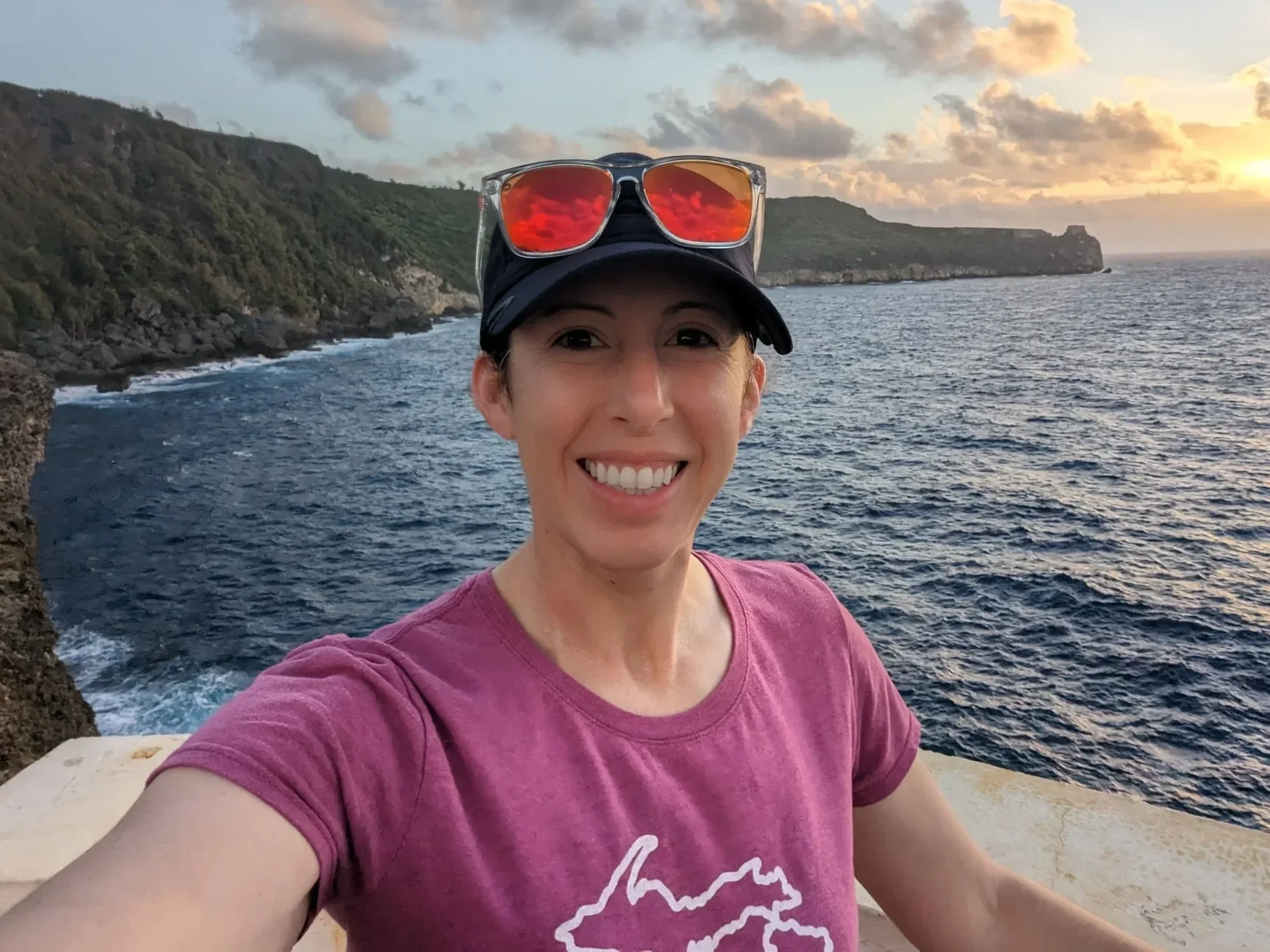 A woman taking a selfie near the coast during sunset, wearing a black cap, sunglasses with red reflective lenses on her head, and a purple t-shirt with a white outline of Michigan on it. The ocean and cliffs are visible in the background.