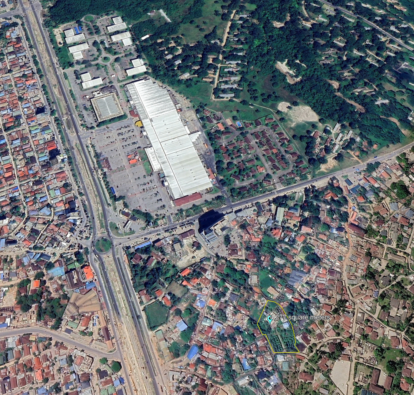 An aerial view of a city neighborhood showing residential houses, a large commercial shopping area with parking lot, and a nearby green park with trees.