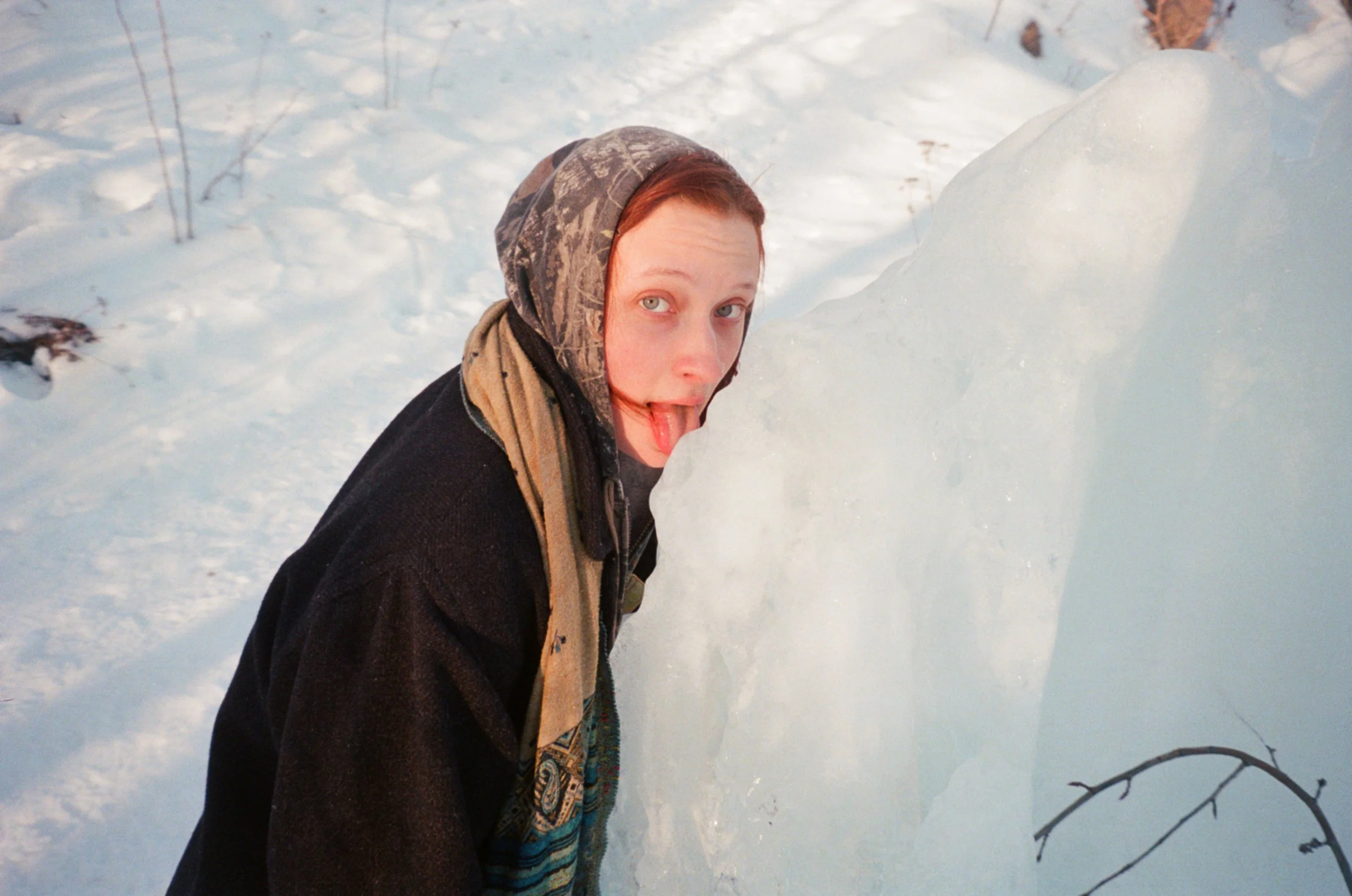 A person with red hair and a hooded jacket sticking out their tongue, standing in a snowy outdoor area next to a snow mound.