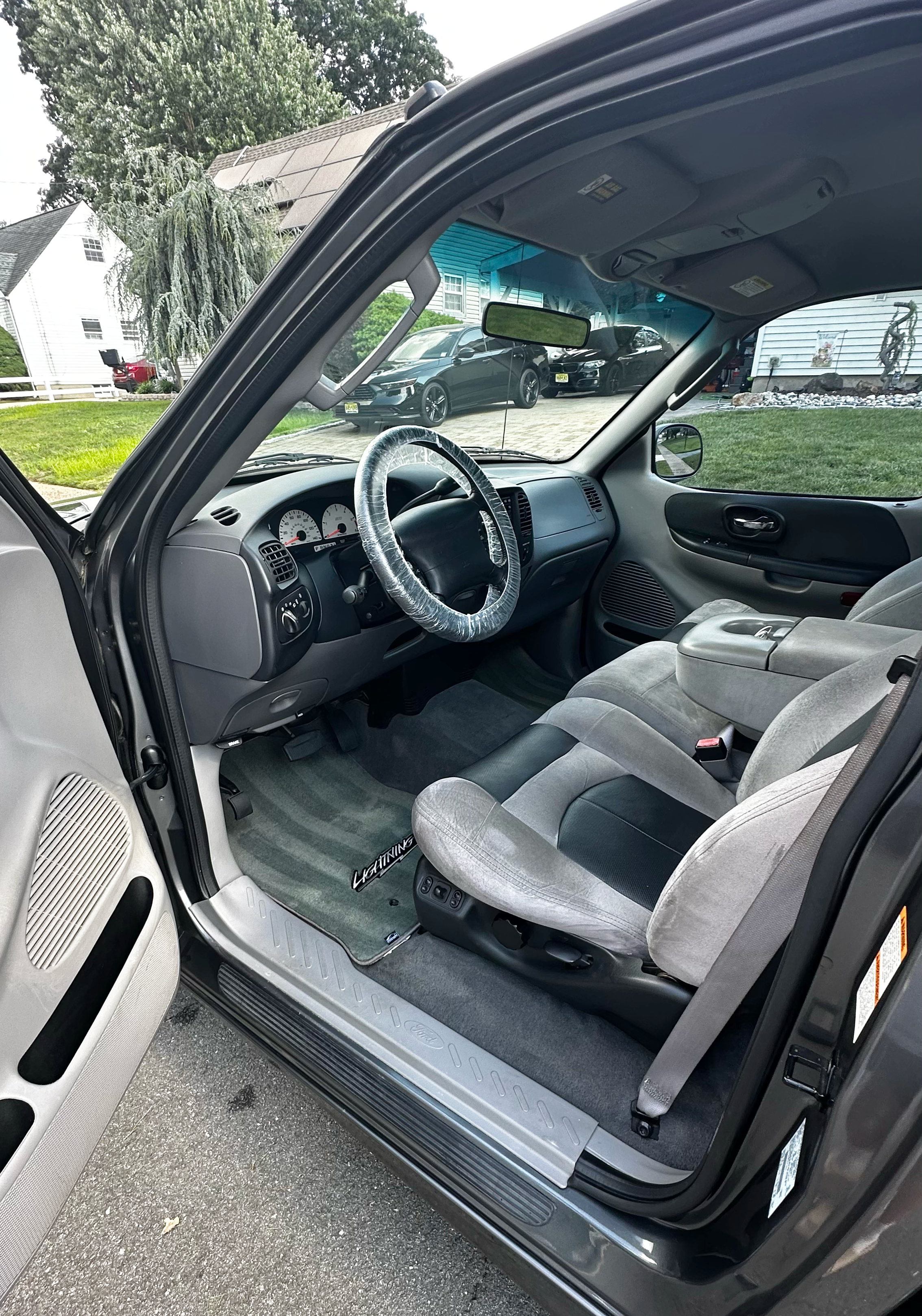 Inside view of a vehicle showing the driver's seat with a plastic cover on the steering wheel, a dashboard, and the front passenger seat. The exterior environment is a driveway with several houses and cars parked.