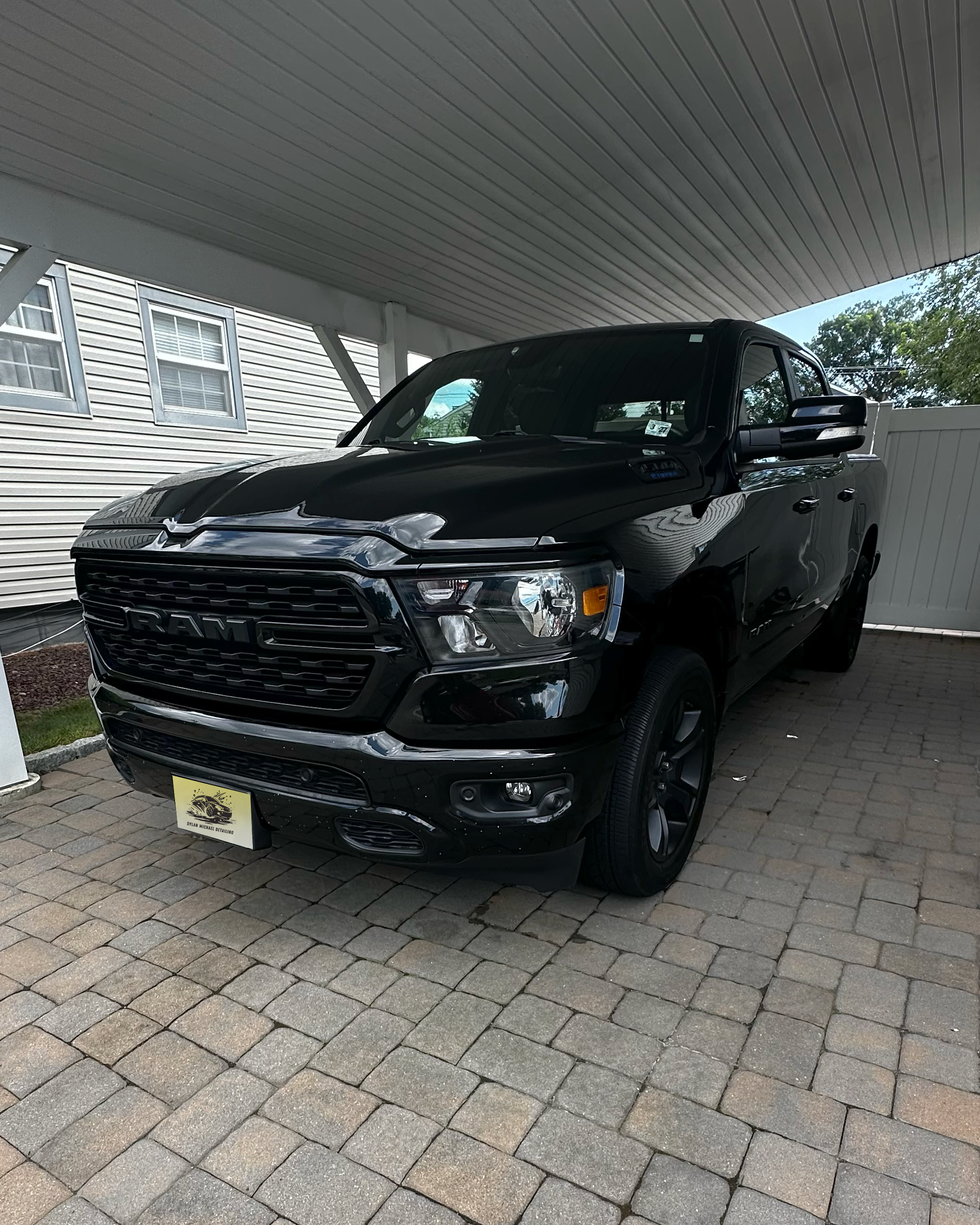 A black RAM pickup truck parked on a paved driveway underneath a porch with a white ceiling. The background includes a white house with three windows and some trees.