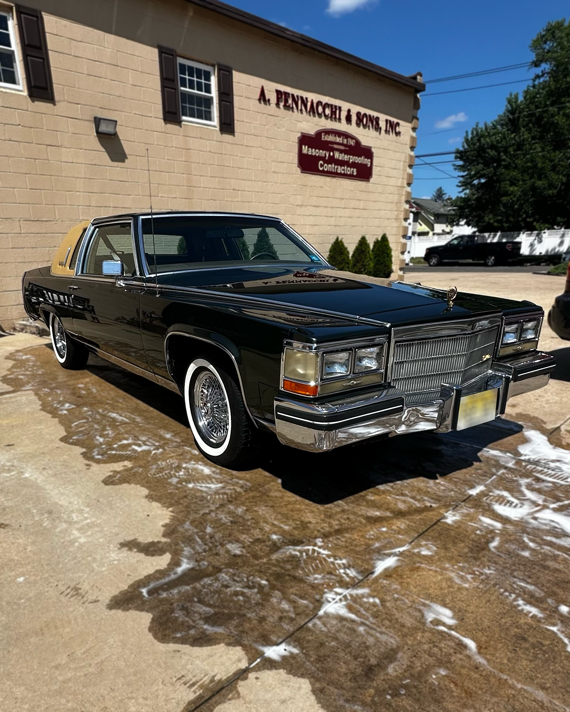 A classic black convertible car with cream-colored interior parked outside a beige brick building with a sign that reads 'A. Pennacchi & Sons, Inc., Established in 1904, Masonry Waterproofing Contractors.' The building has two windows with brown shut