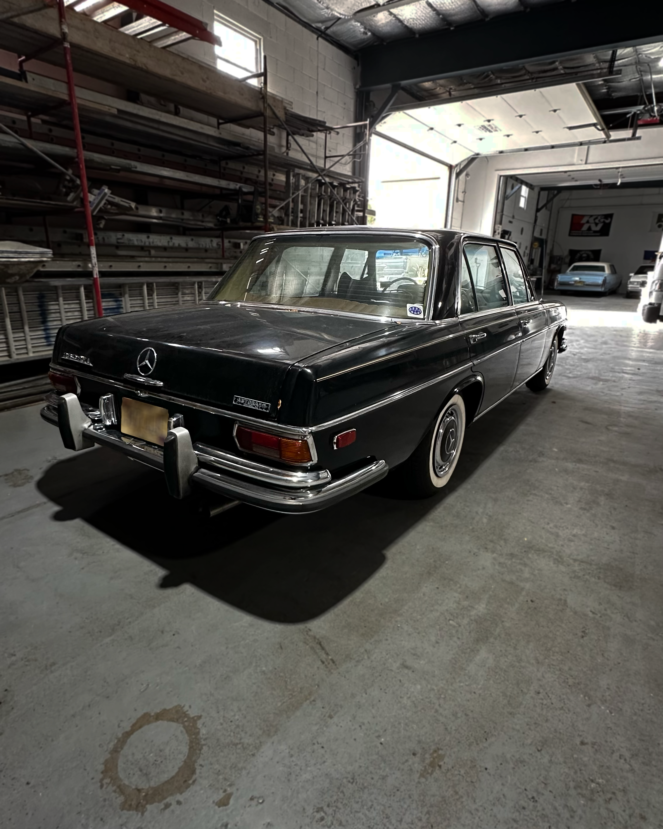 A black classic Mercedes-Benz car parked inside a garage with an open garage door. The garage has shelves and car parts along the walls, and other cars are visible in the background.