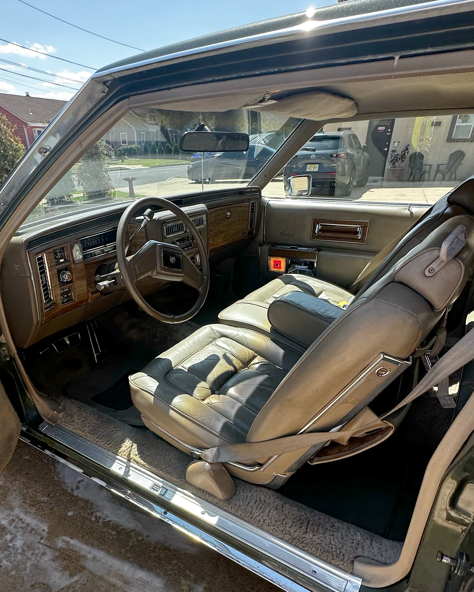 Inside a vintage car showing the dashboard with wood paneling, a steering wheel, and a front bench seat with older upholstery. The view through the windshield reveals parked cars and houses on a sunny residential street.