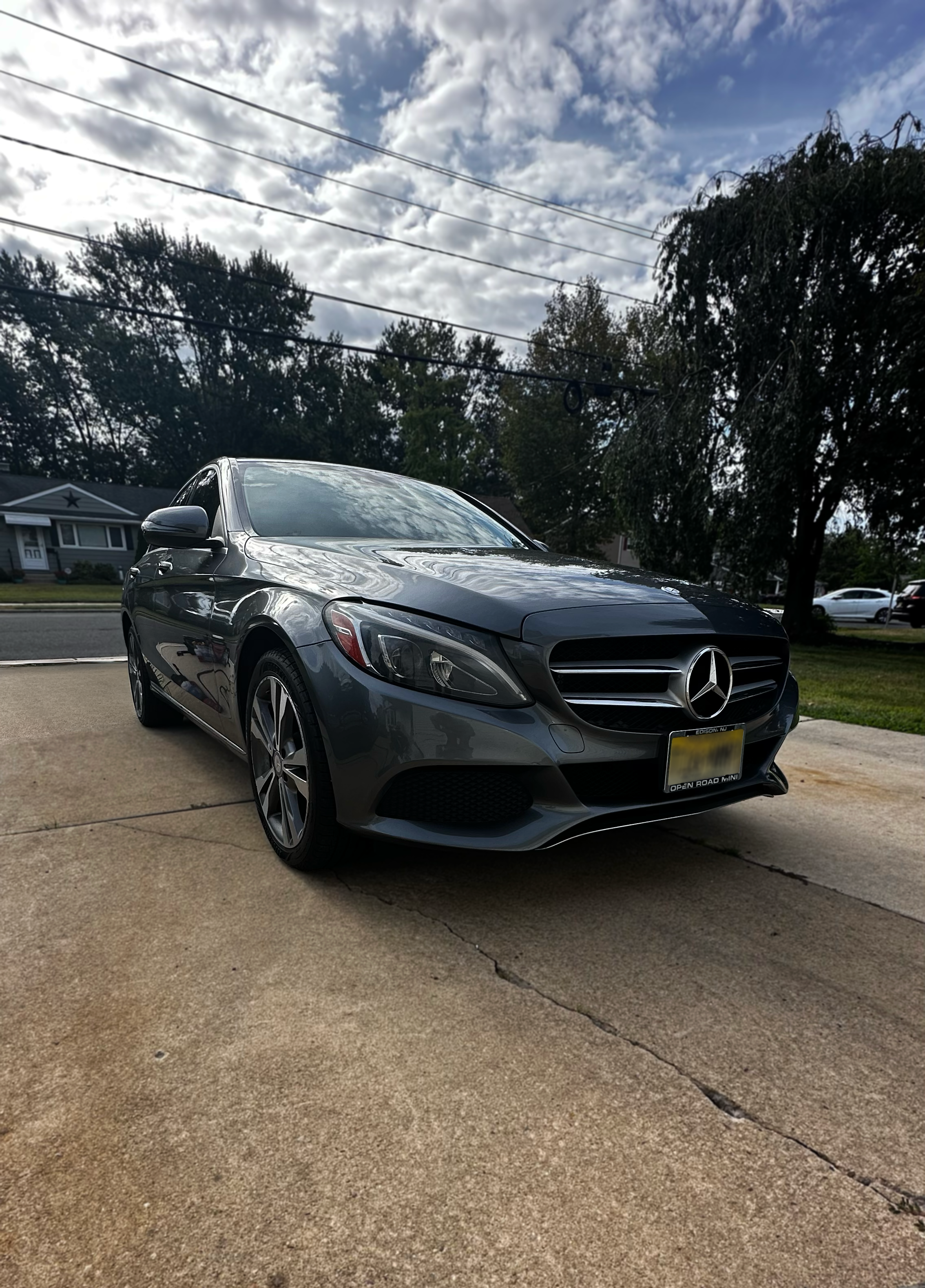A shiny gray Mercedes-Benz parked on a concrete driveway in front of a suburban house with trees, a cloudy sky, and power lines overhead.