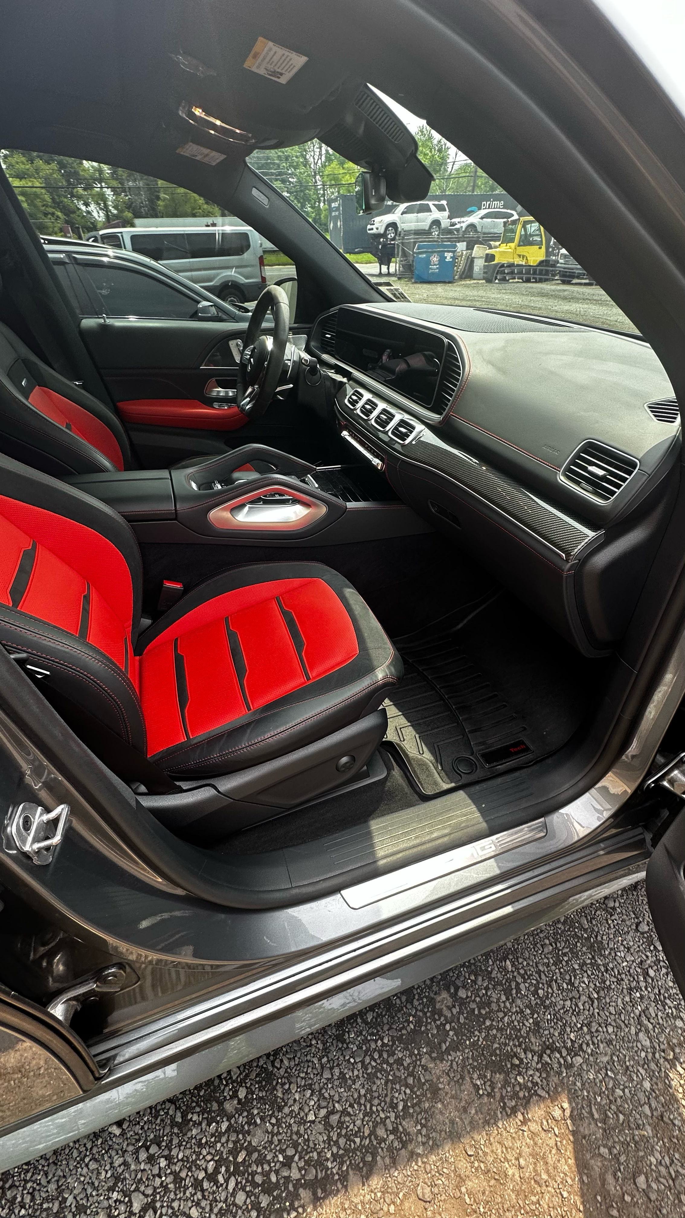 The interior of a modern car with black and red racing seats, dashboard, and steering wheel, with a background showing an outdoor parking lot.