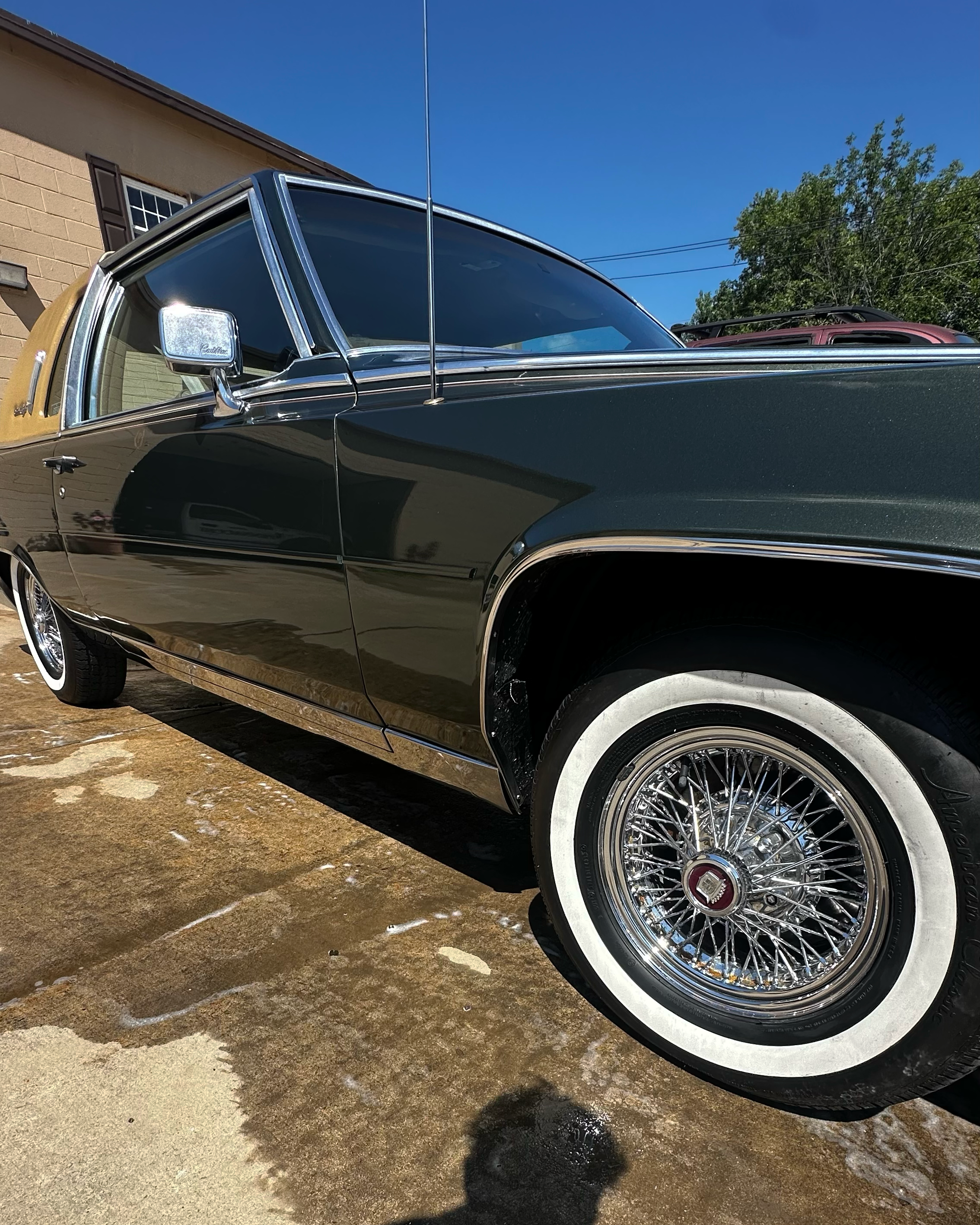 Close-up of a vintage black car with whitewall tires, chrome wire wheels, and chrome accents parked outside on a sunny day, with a house and a tree in the background.