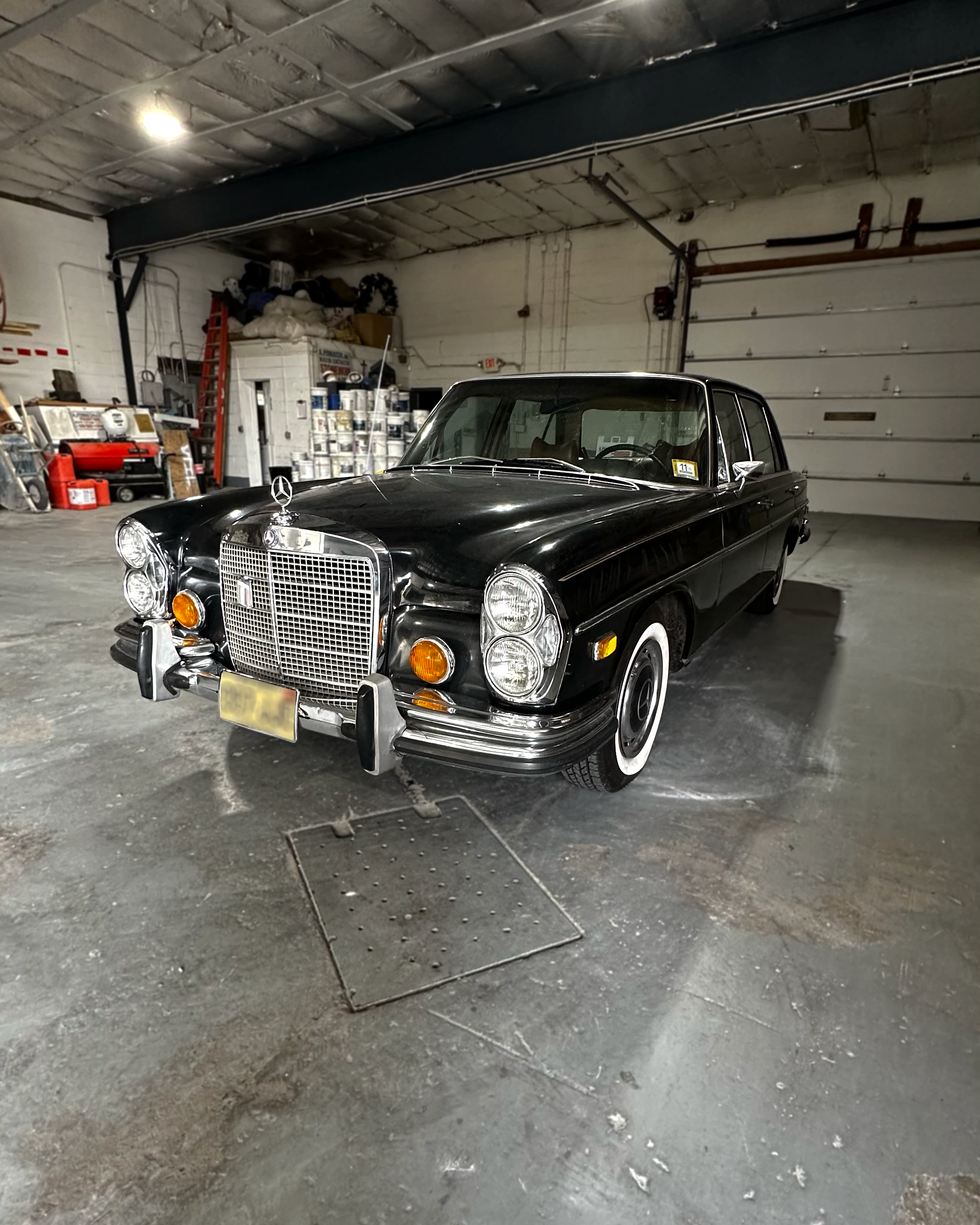 A black vintage Mercedes-Benz car parked inside a garage with a concrete floor, overhead lights, and various storage shelves and items in the background.