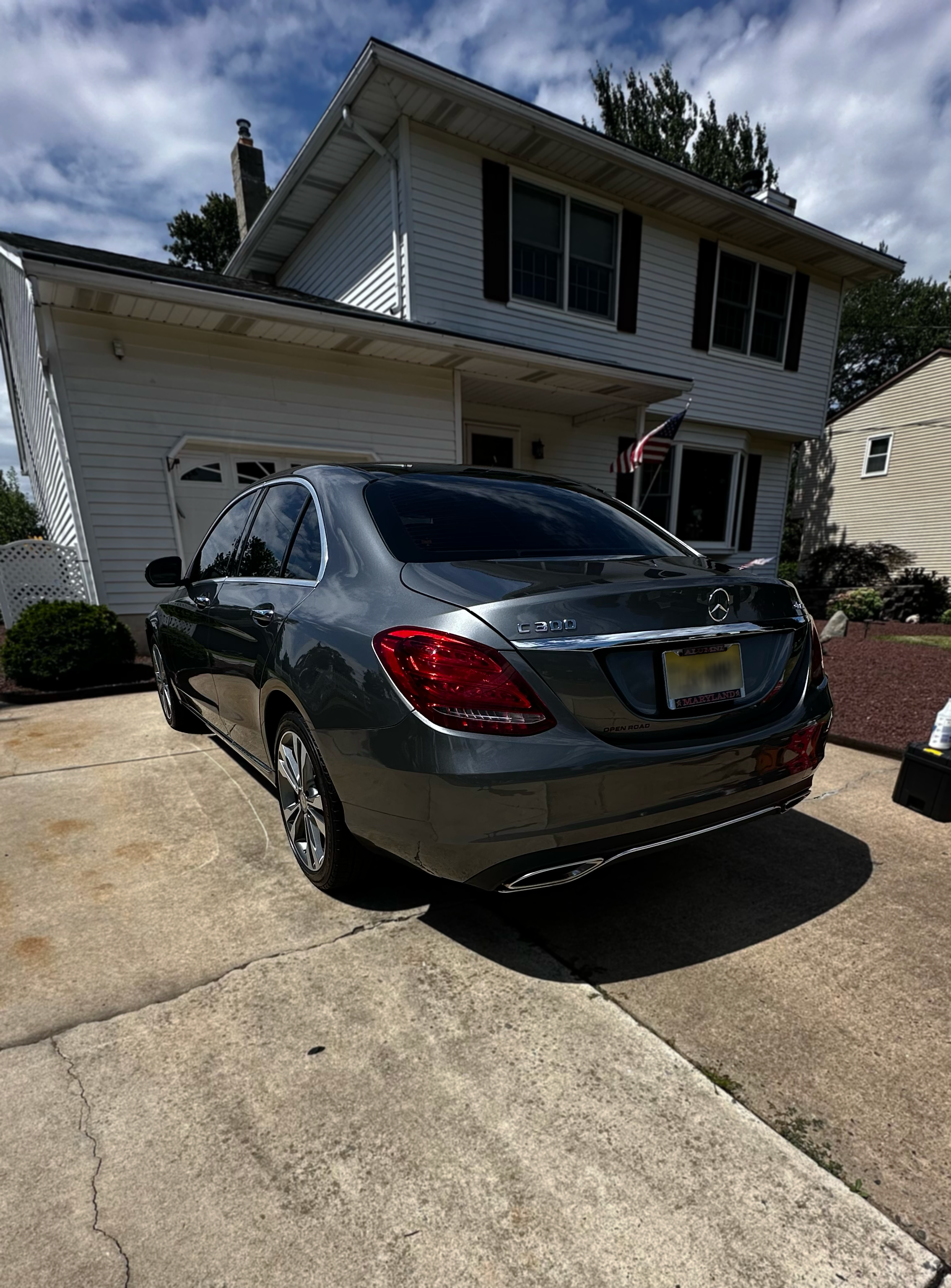A black Mercedes-Benz C300 parked in a driveway in front of a white two-story house with black shutters and an American flag hanging outside.