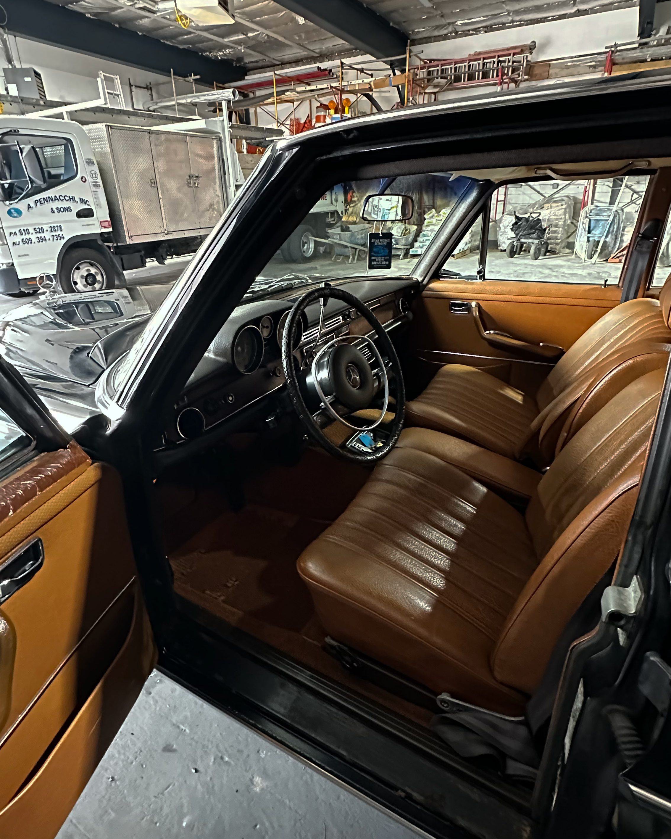 Inside a vintage car with tan leather seats, viewing the dashboard and steering wheel through the open driver's side door, with a workshop or garage visible in the background.
