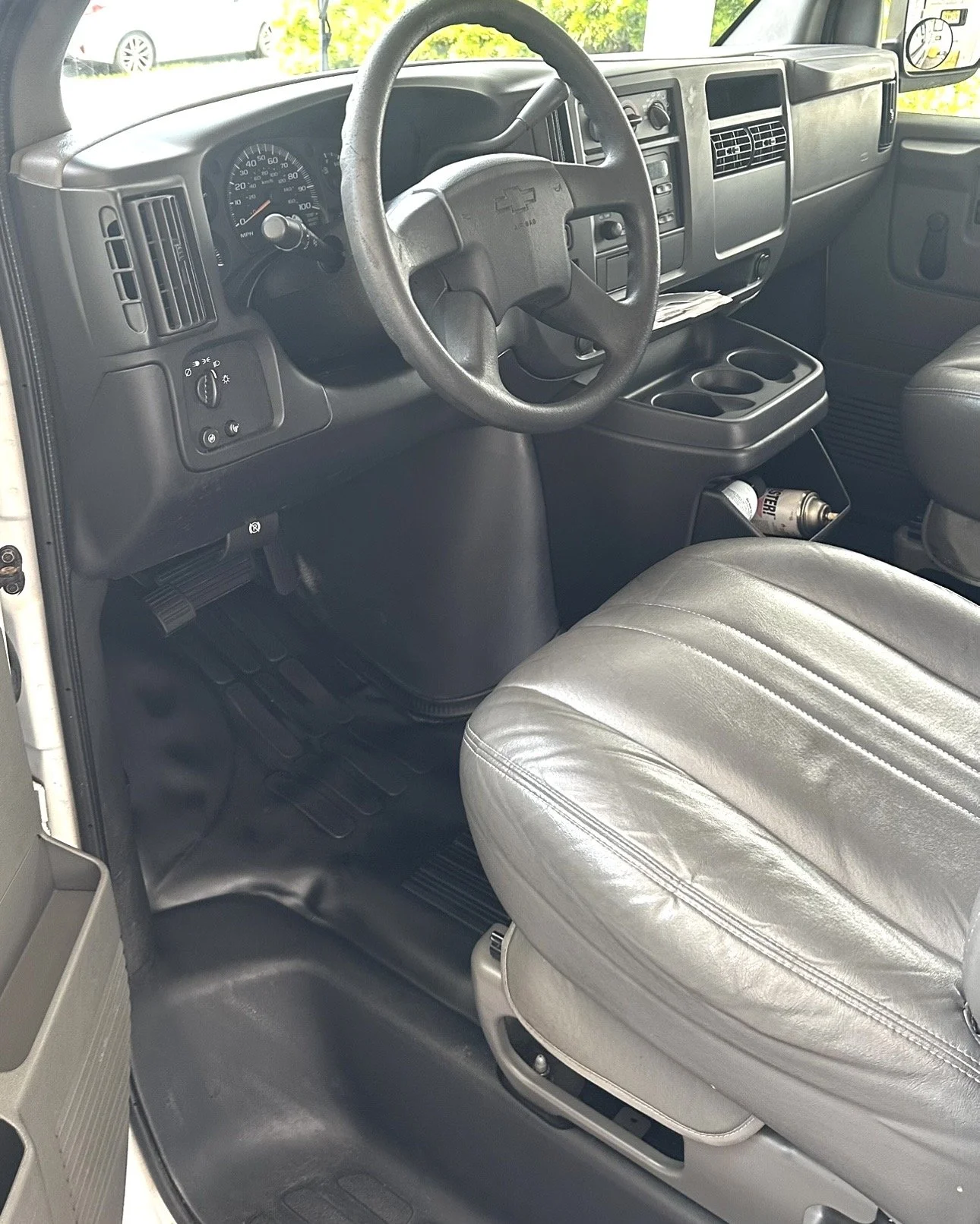 Inside the cab of a commercial truck, featuring a steering wheel with Chevrolet logo, dashboard with gauges, a fire extinguisher under the seat, and a cup holder next to the seat.