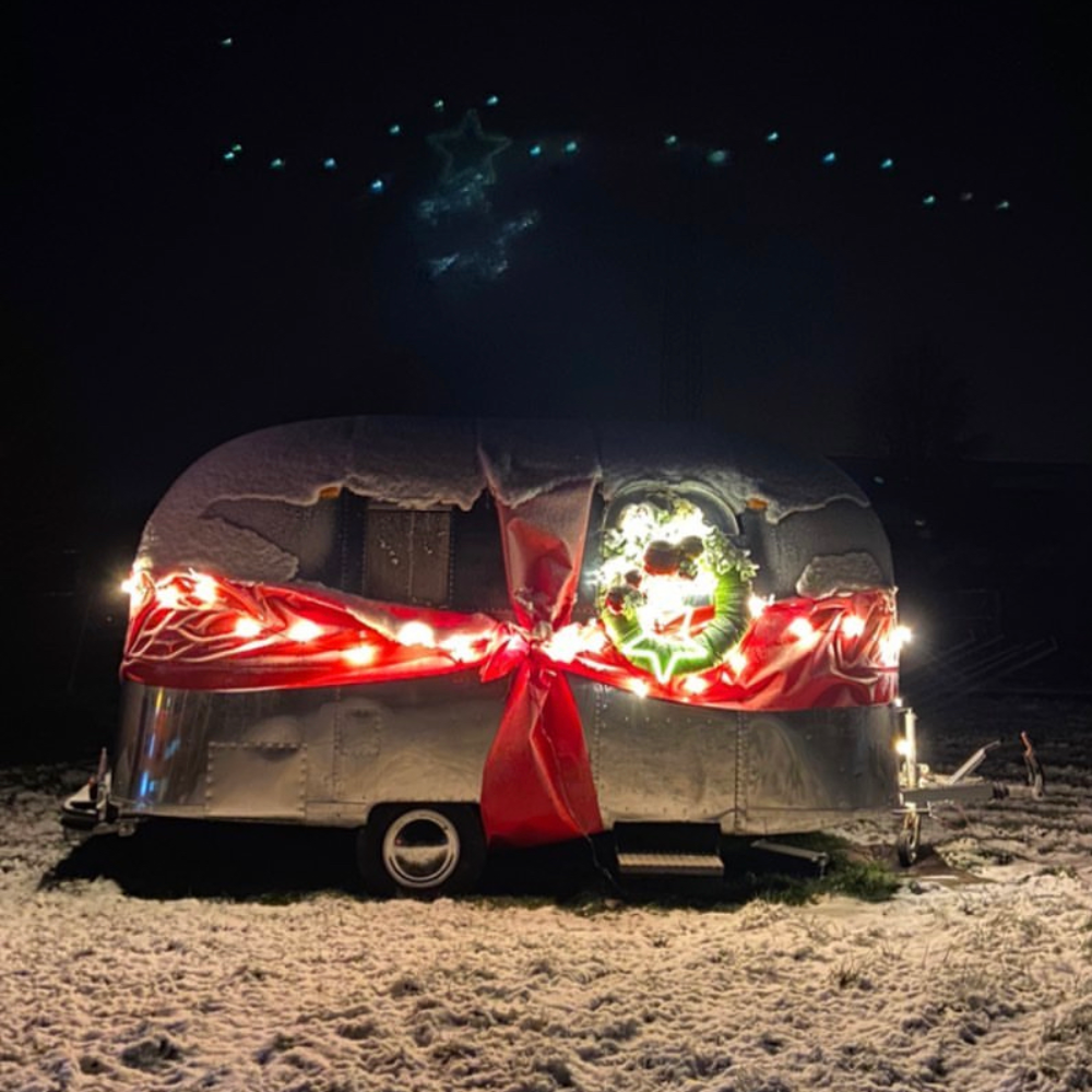 Ein Airstream im Freien, bedeckt mit Schnee. Der Anhänger ist mit einem großen roten Band und einer beleuchteten Girlande dekoriert. Im Hintergrund ist ein Sternenhimmel zu erkennen.