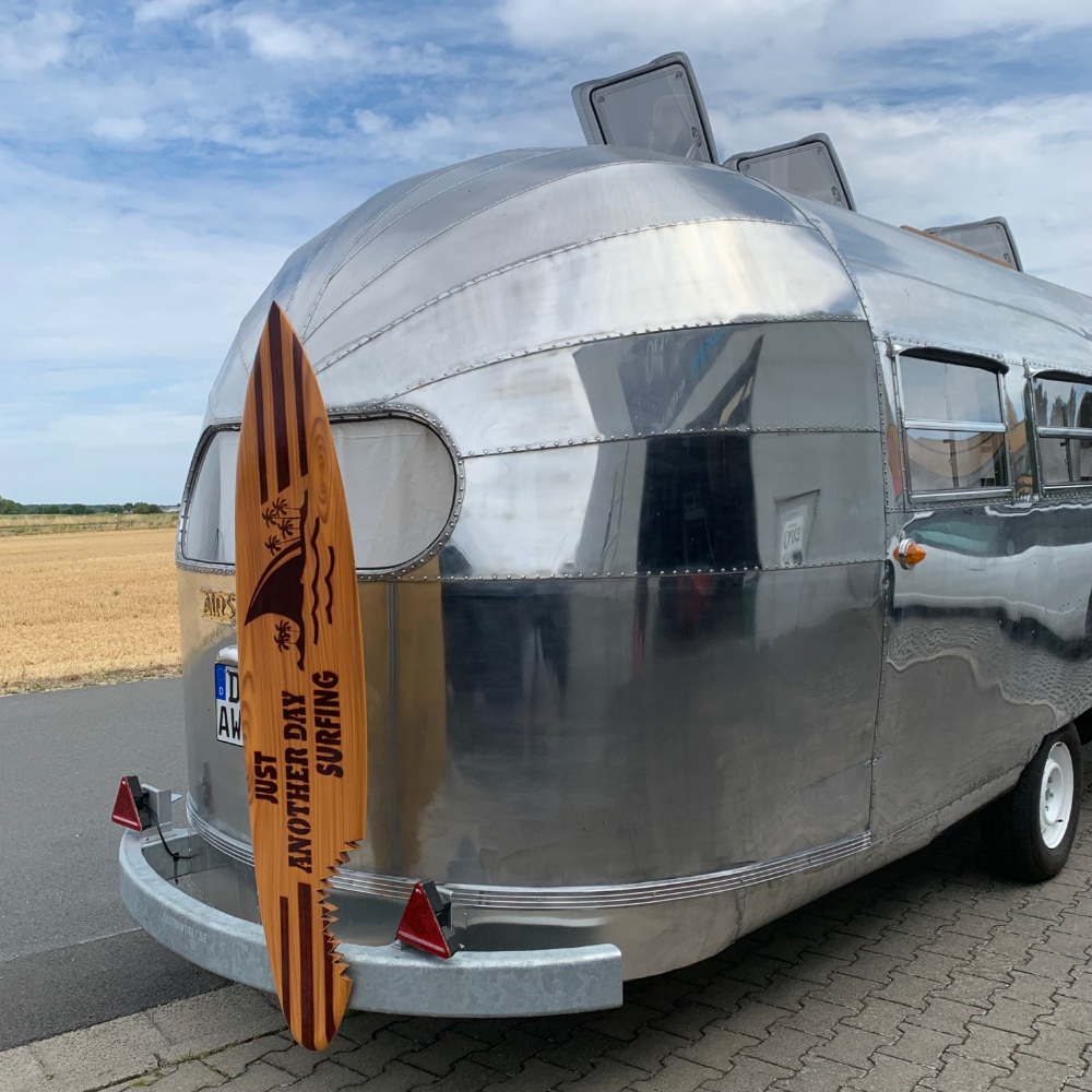 Silberner Wohnwagen mit einem Holzschild an der Rückseite, auf dem 'Just Another Day Surfing' steht, auf einem Parkplatz mit einem Feld im Hintergrund.