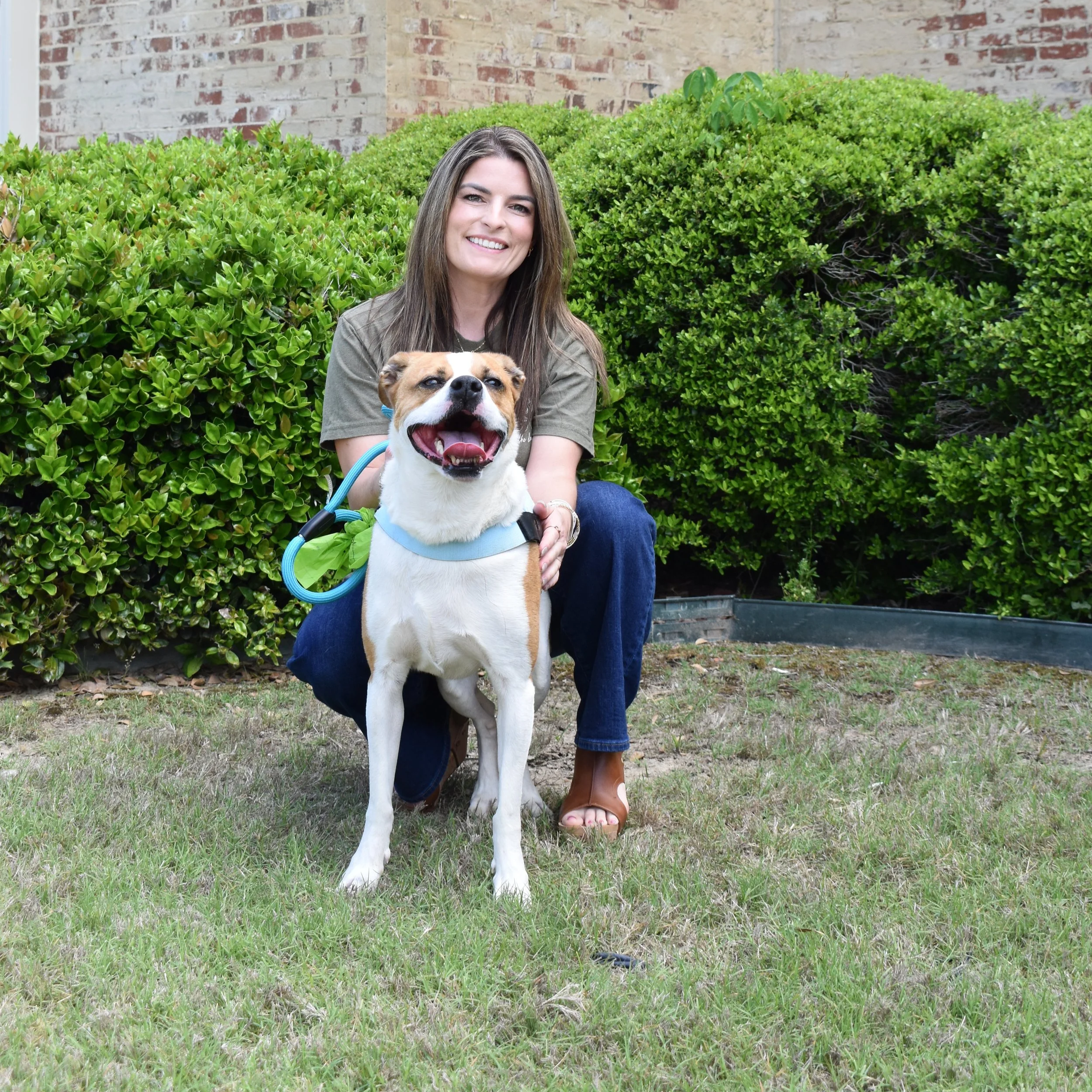 A woman standing in front of a brick wall, wearing an olive-green sweatshirt with a small graphic of a dog on the left chest.