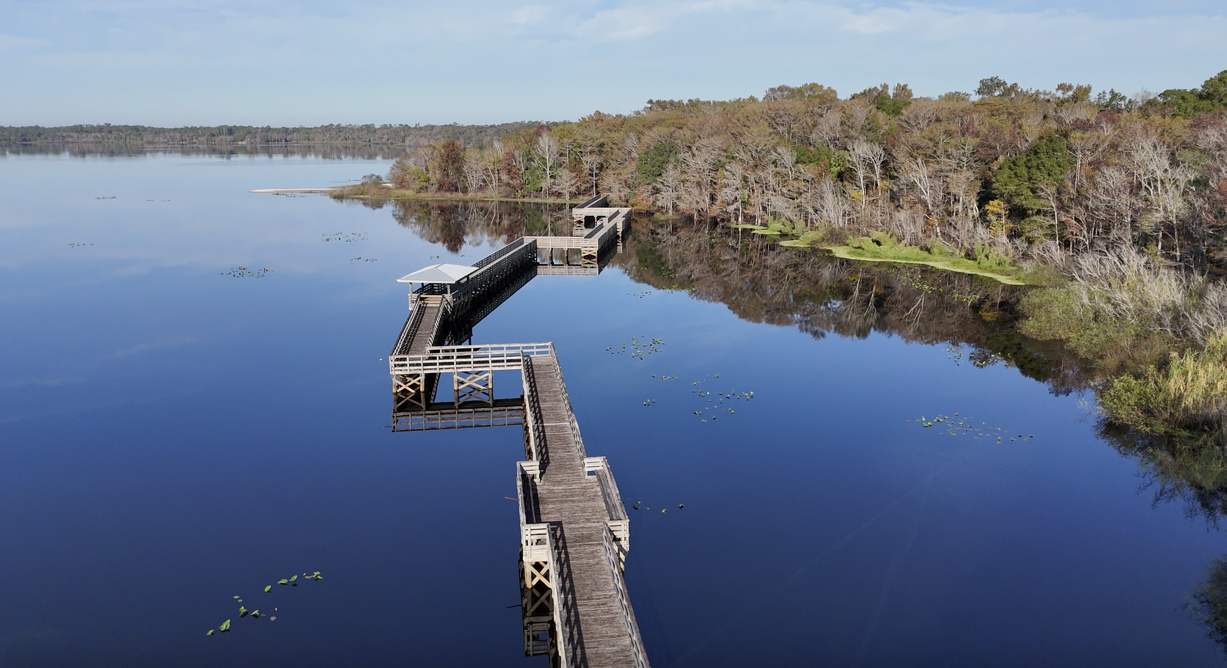 A wooden pier extending over calm water with a small covered section at the end, surrounded by trees with autumn foliage, reflecting on the water's surface under a clear sky. Pier built by Orlando based contractor specializing in docks and seawalls.