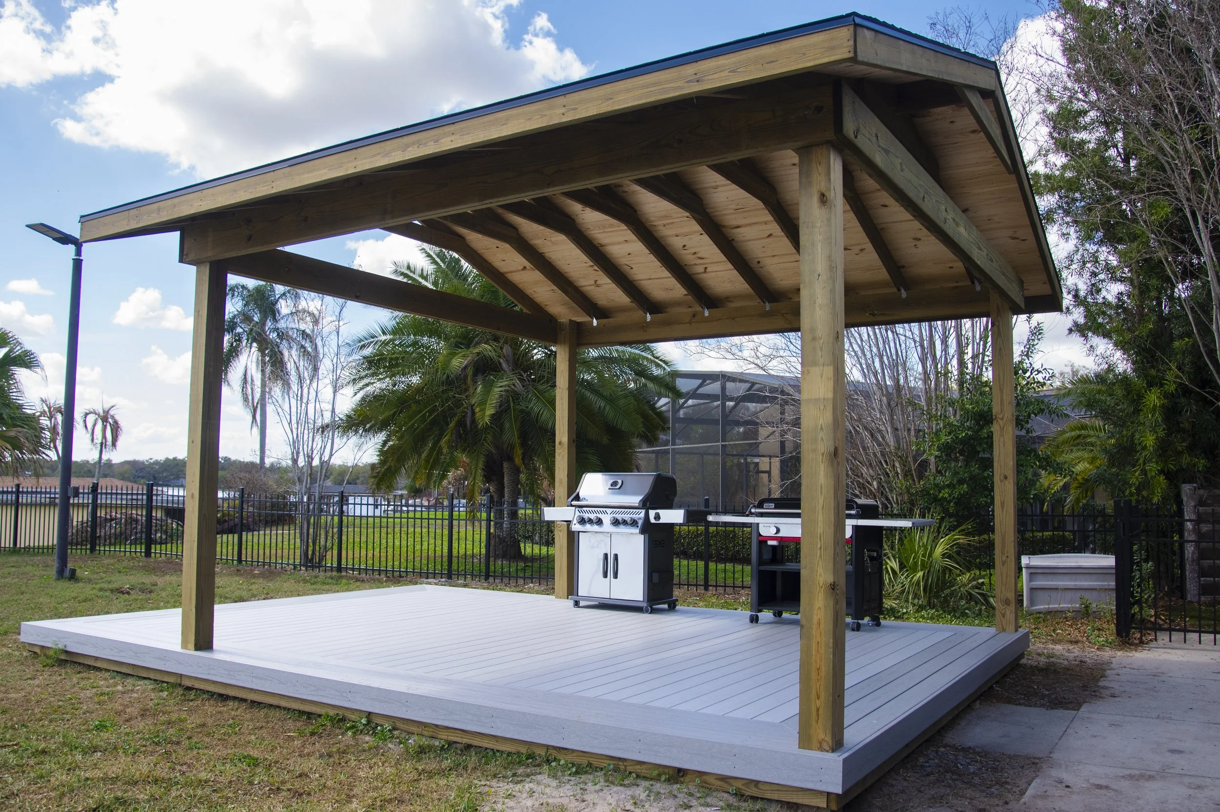 A wooden outdoor deck with a roof, supported by wooden posts, with a gas grill and outdoor furniture, surrounded by a fenced yard with trees and a clear sky.