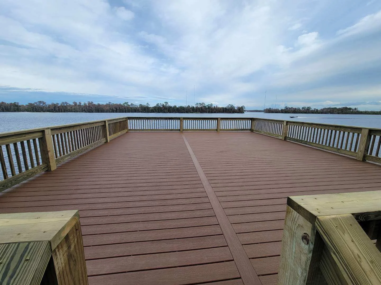Empty wooden deck overlooking a body of water with trees in the distance under a partly cloudy sky. Two story deck with composite decking.
