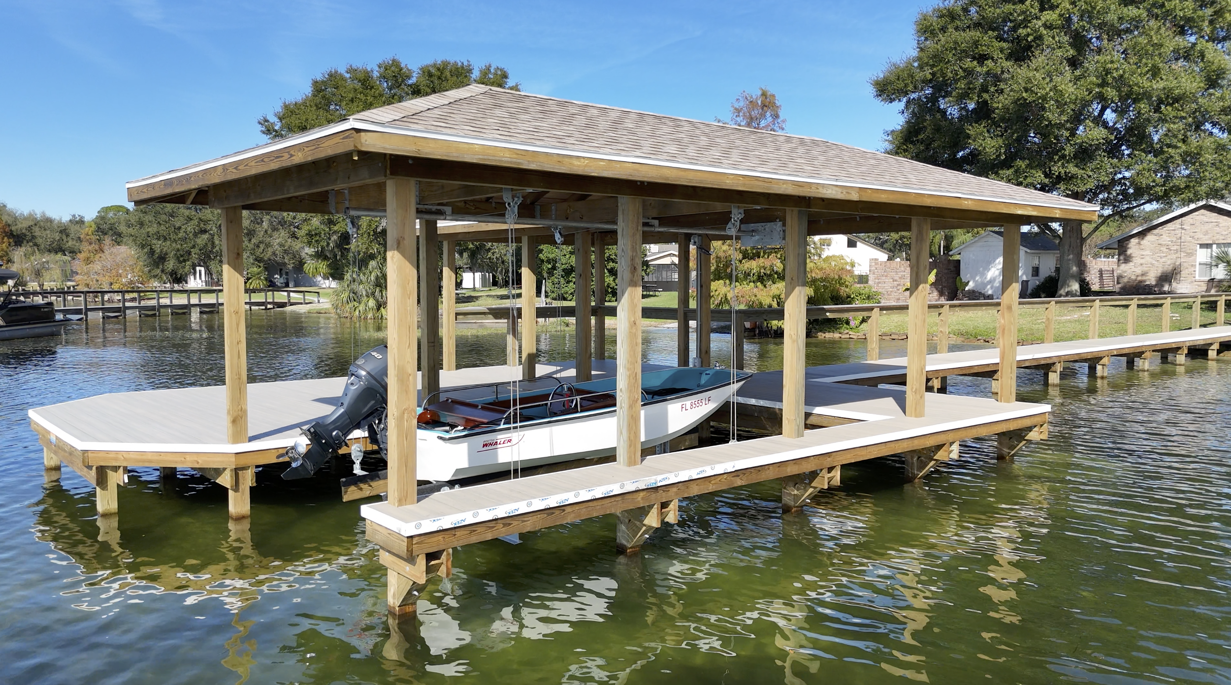 Waterfront boat dock with boat and motor, surrounded by trees and houses on a clear day. Built by an expert dock builder out of marine grade materials.