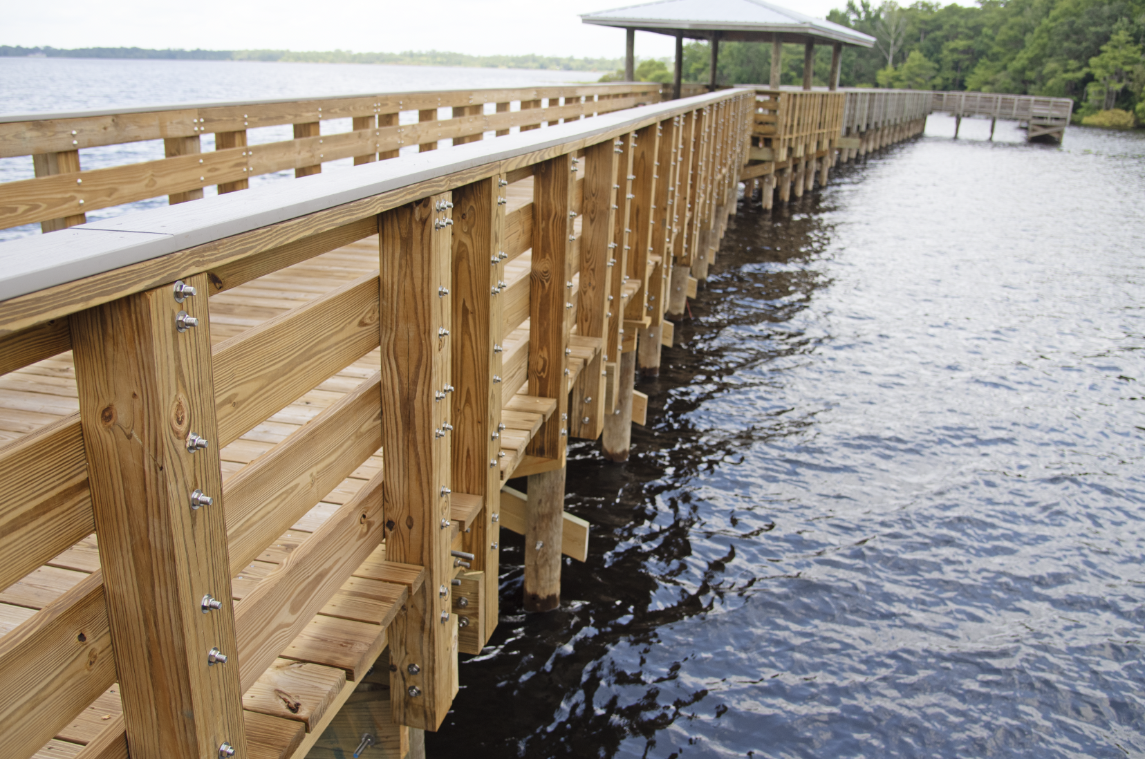 Wooden pier extends over a body of water, with trees in the background and a shelter at the end of the pier.