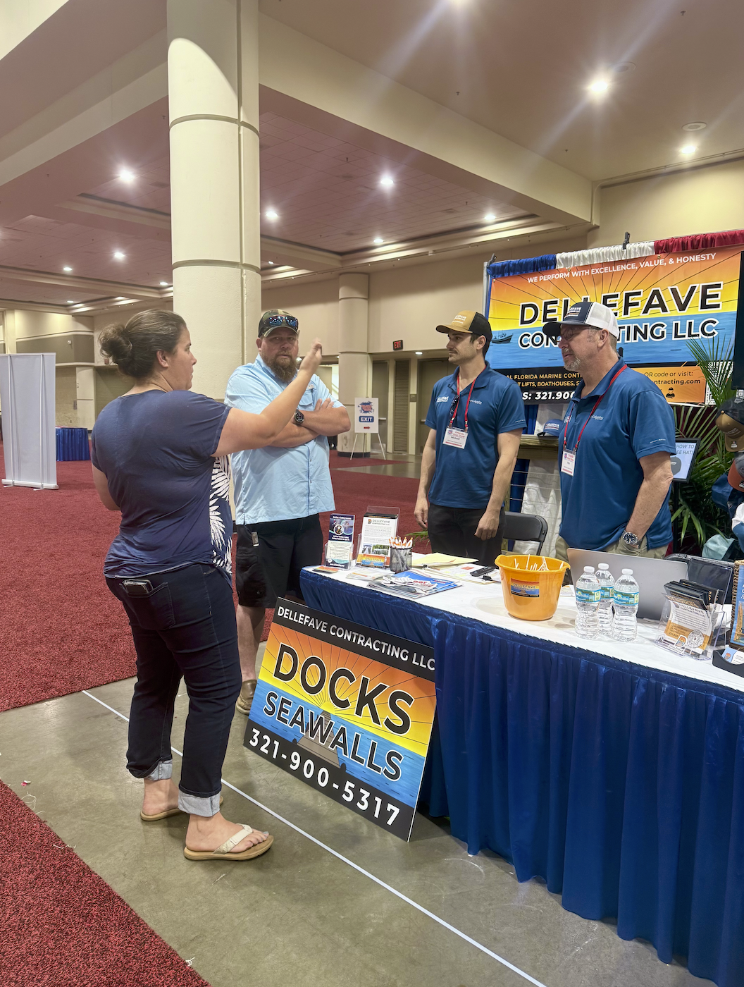 A woman is talking to three men at a booth for Dellefave Contracting LLC, which specializes in docks and seawalls. The booth has a large sign with the company's name, contact information, and a display of brochures, pens, and bottled water.