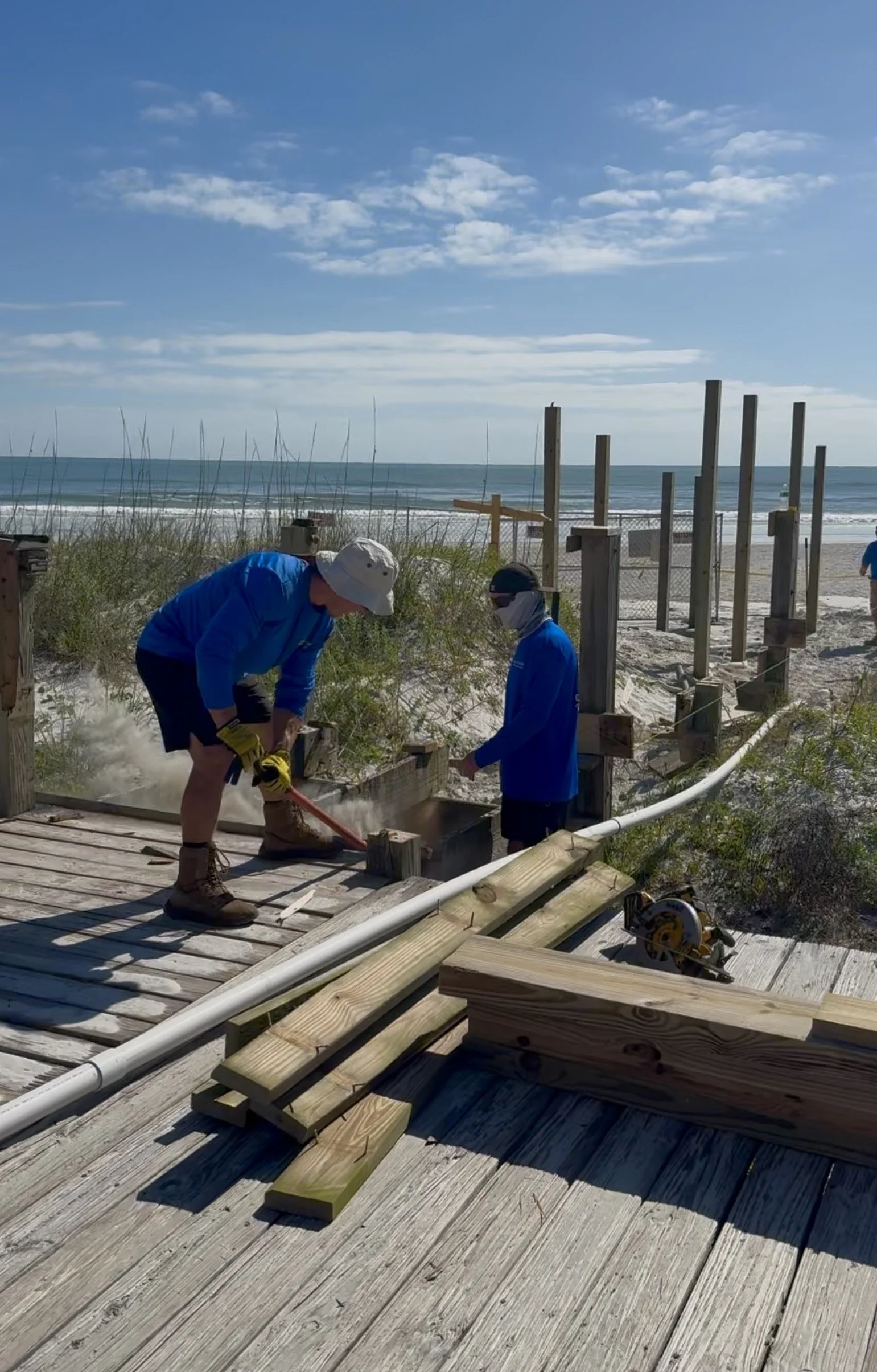 Two people working on building a boardwalk on the beach. One person is using a power tool while the other is assisting. There are wooden planks and construction tools on the deck, with the ocean and cloudy sky in the background.