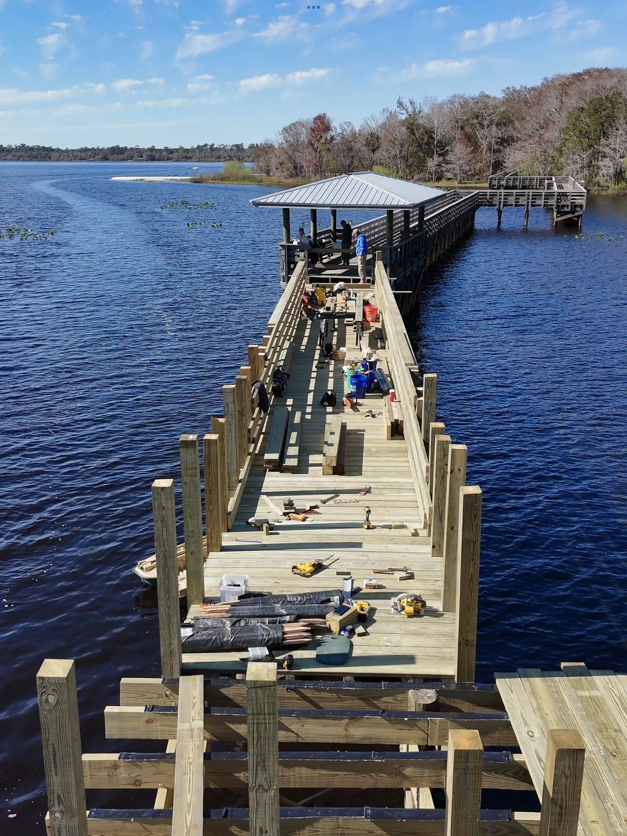 Expert dock builders building a wooden pier or dock over water, with tools and supplies spread out on the planks, trees in the background.