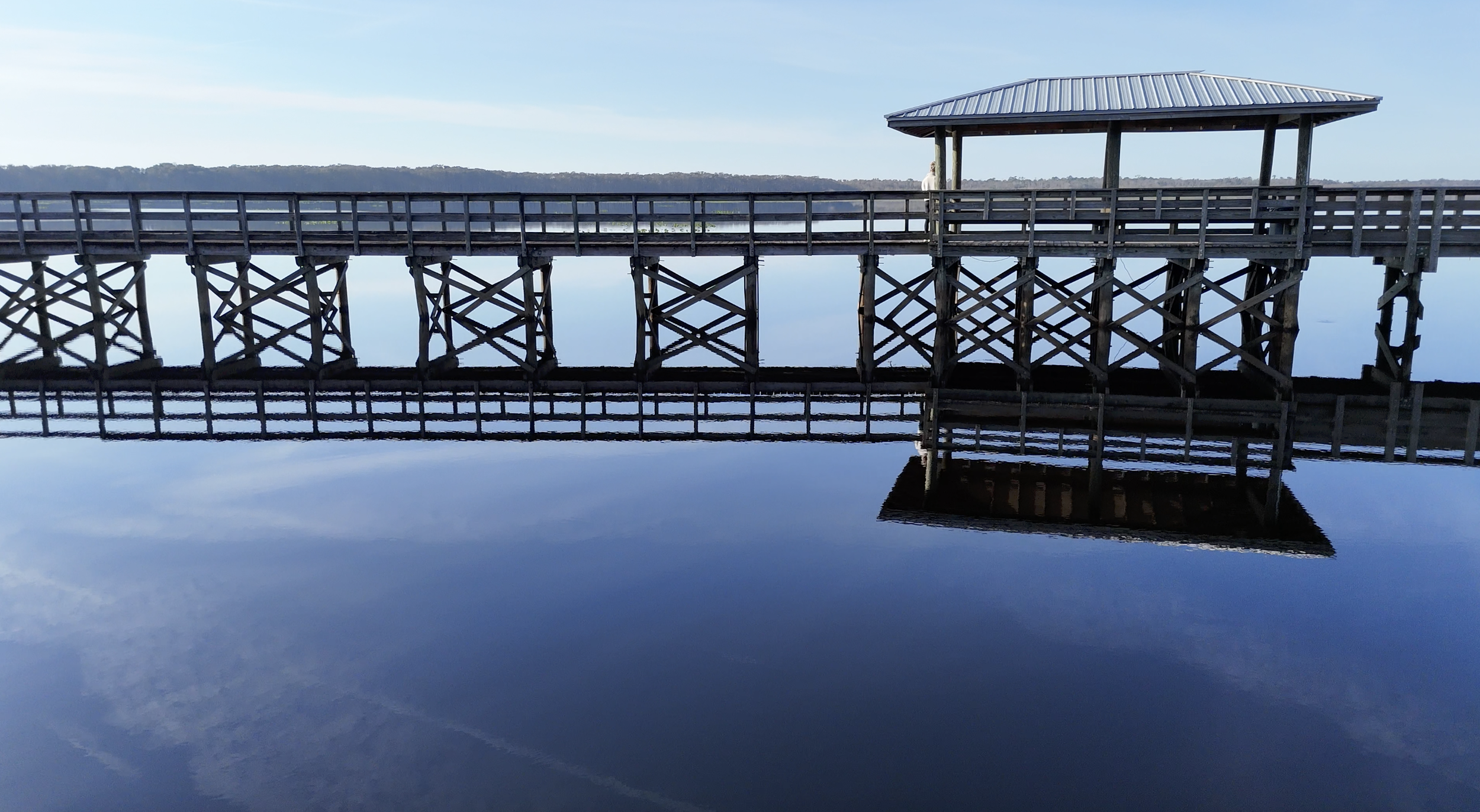 A wooden pier extending over calm water with a small pavilion at the end, reflecting in the water below, and a person standing under the pavilion.