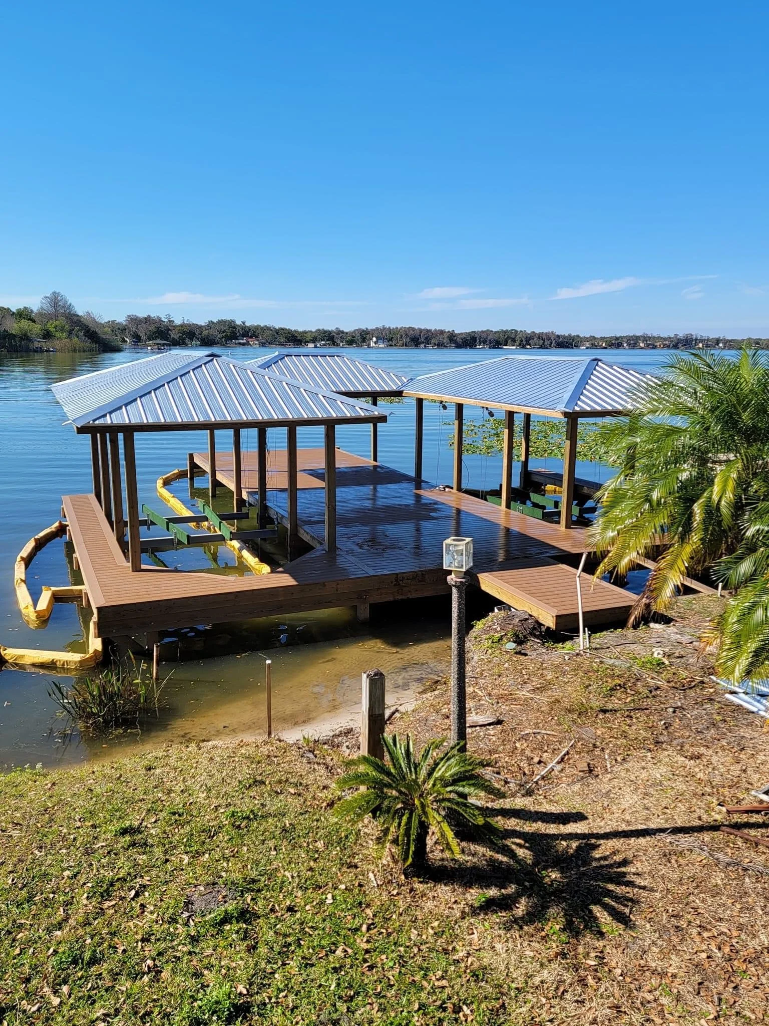 A waterside dock with a metal roof, extending into a calm lake, surrounded by greenery and small plants, with trees in the distance and a clear blue sky.