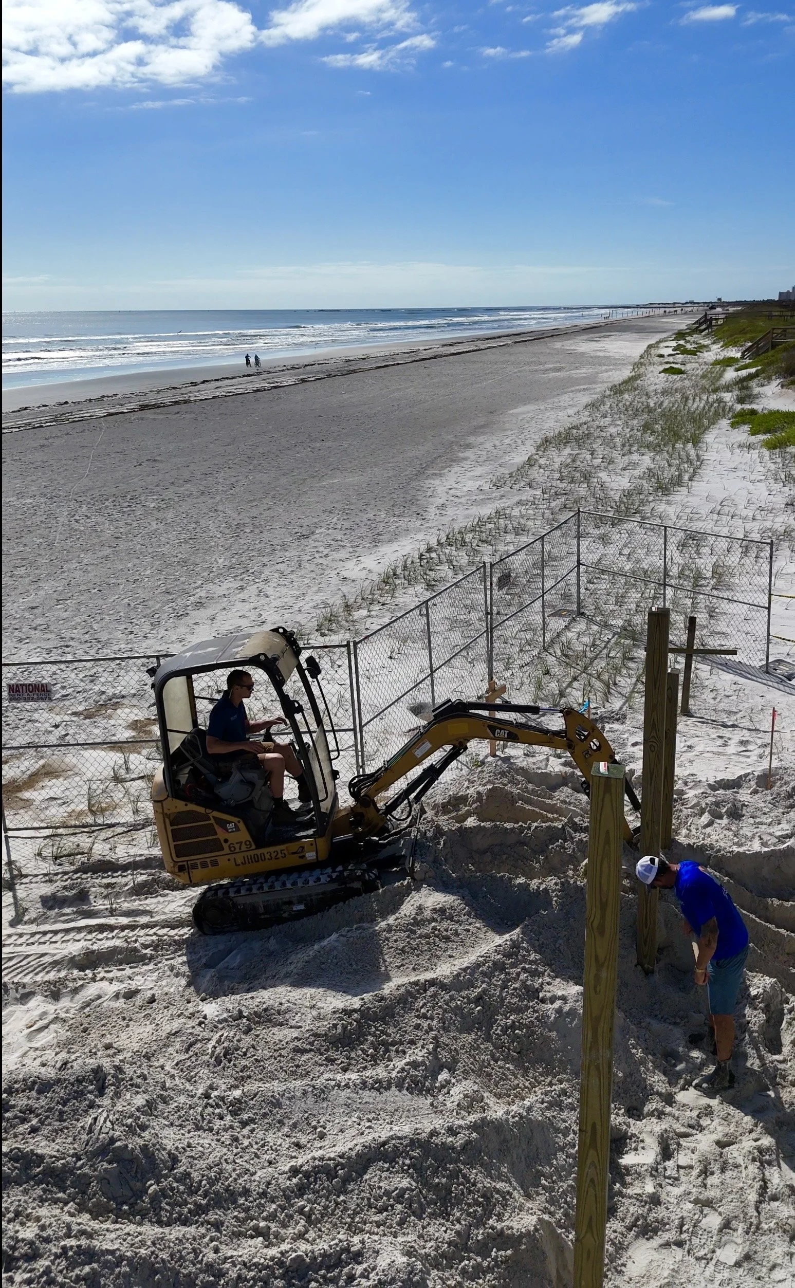 marine constractors workers building a wooden fence on a sandy beach, with a person operating a small excavator nearby and a couple walking along the shoreline in the background.