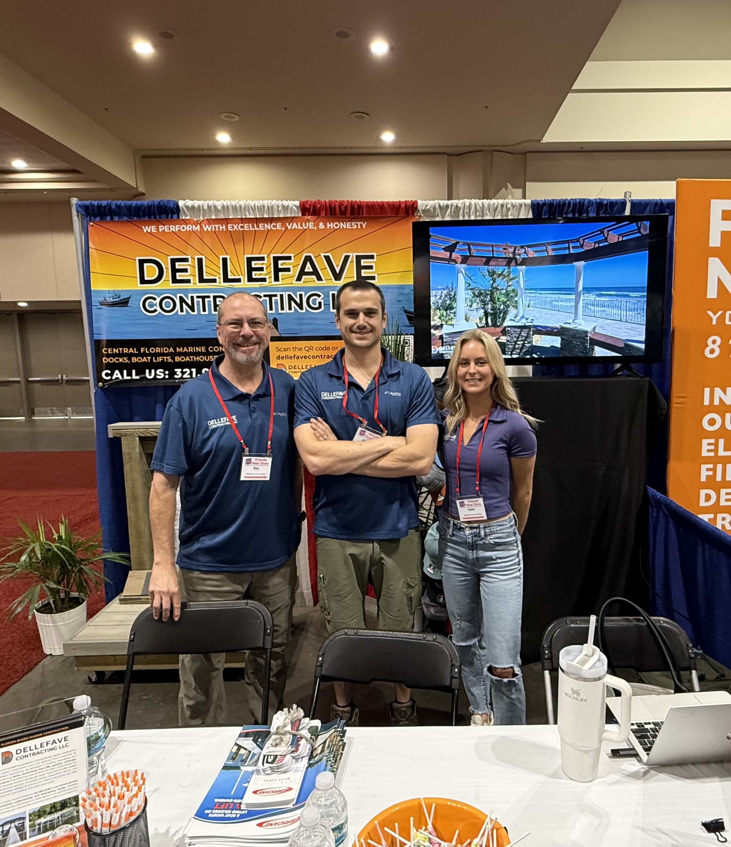 Three people standing in front of a trade show booth for Dellefave Contracting LLC, with a banner and a large screen behind them. The booth displays promotional materials on a table.