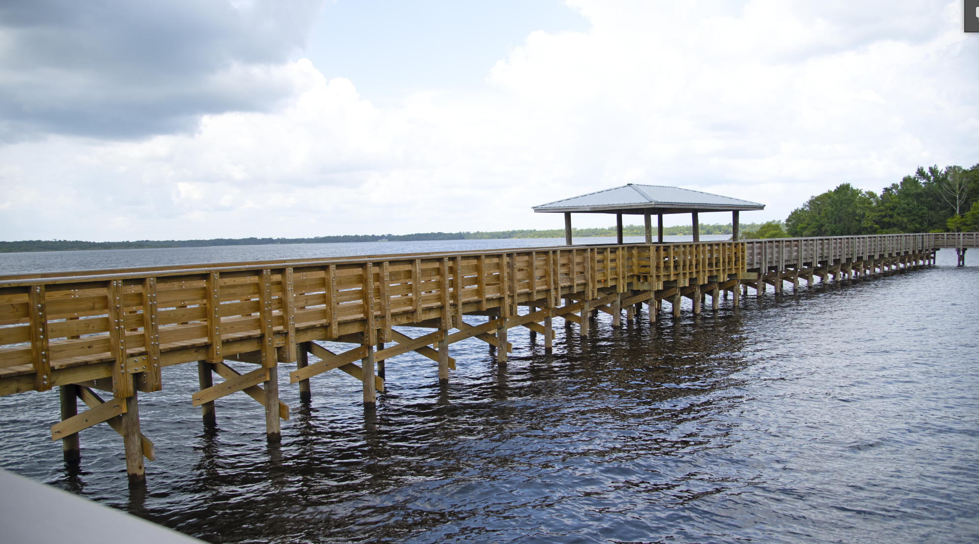 Wooden pier extending over a body of water with a small pavilion at the end, surrounded by trees and clouds in the sky.