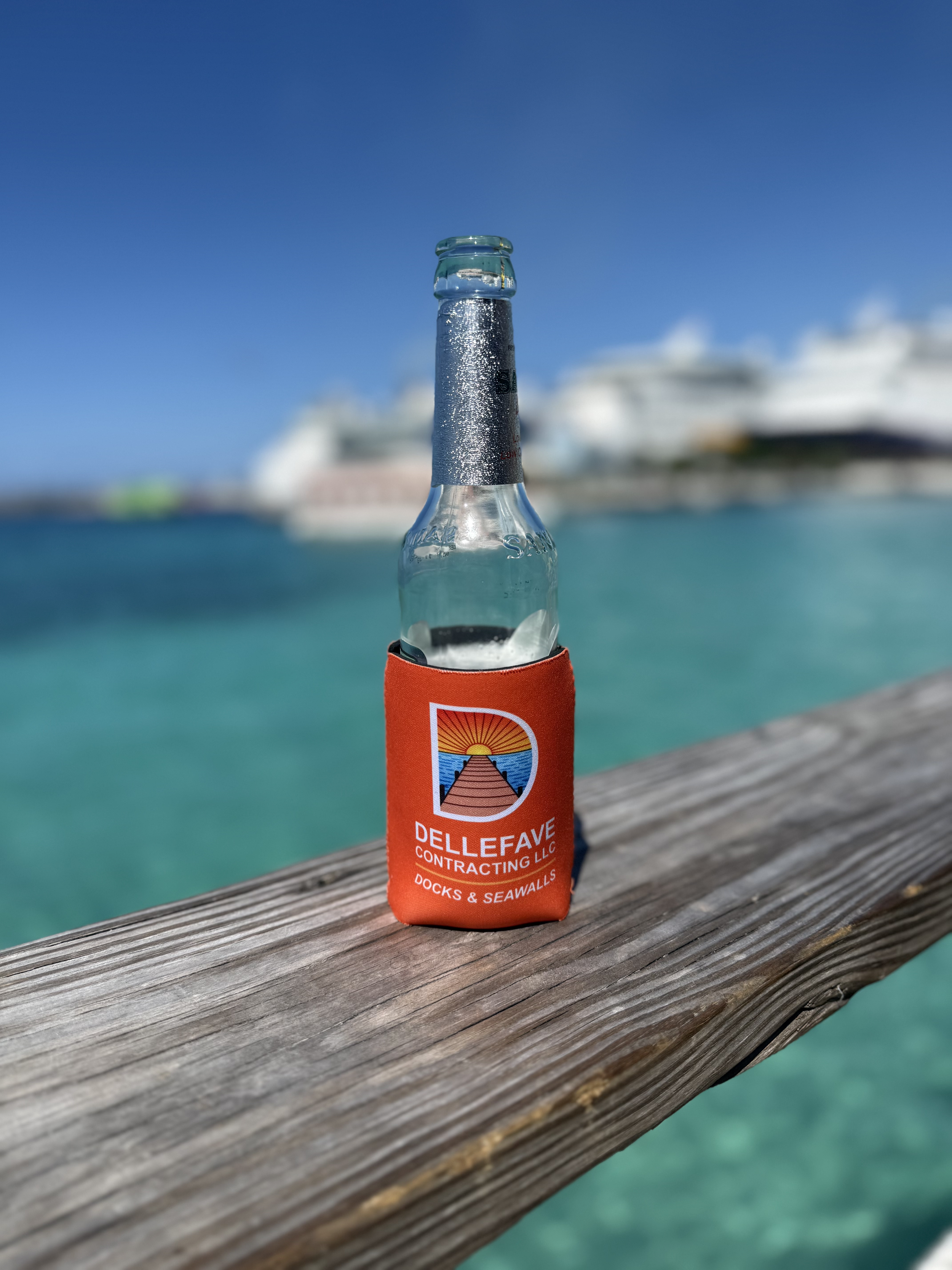 A glass bottle with a 'Dellefave Contracting LLC' sleeve on a wooden dock, with cruise ships and a blue sky in the background.